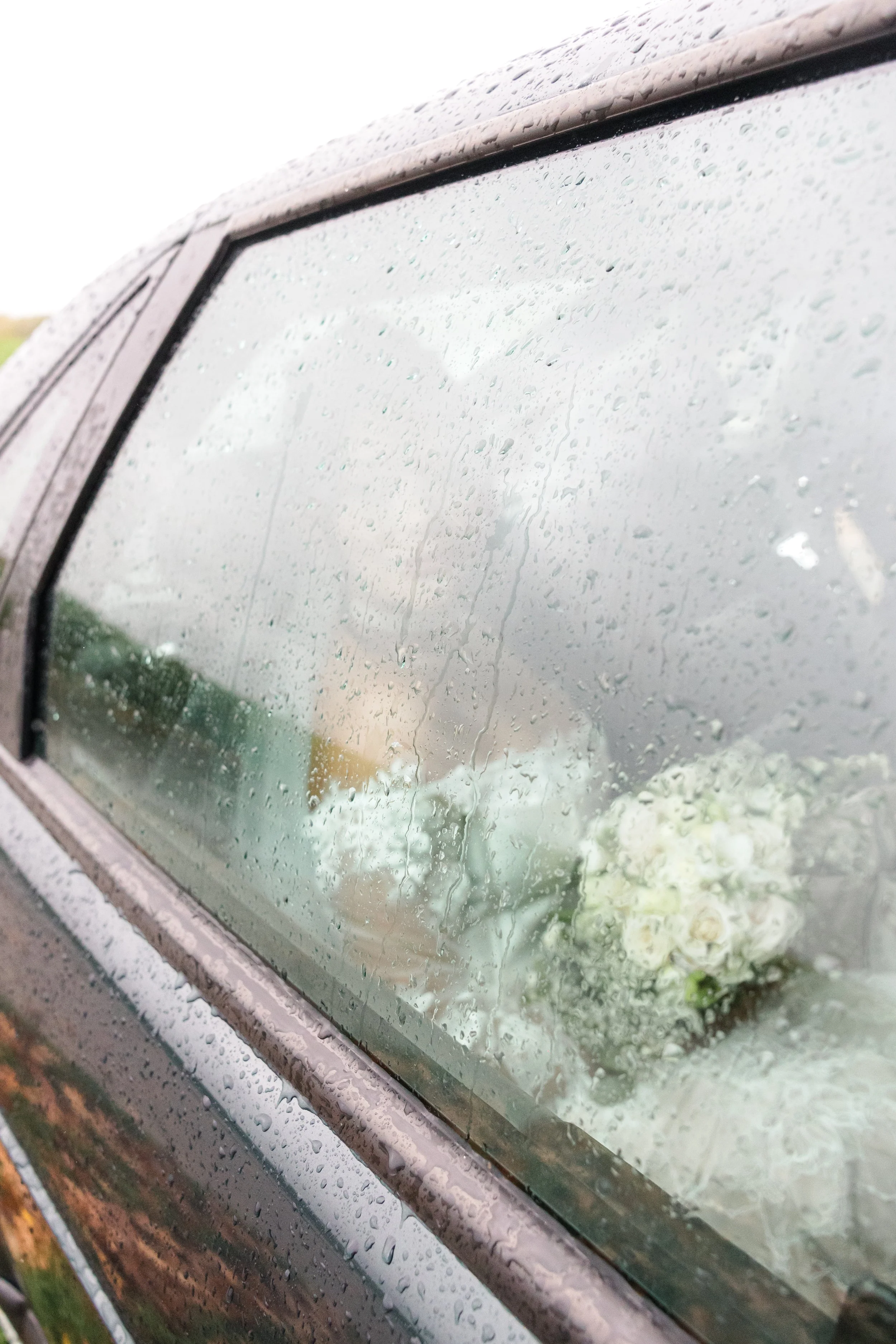 Close-up of a rain-covered car window with droplets of water, and a bouquet of white flowers visible on the seat inside.