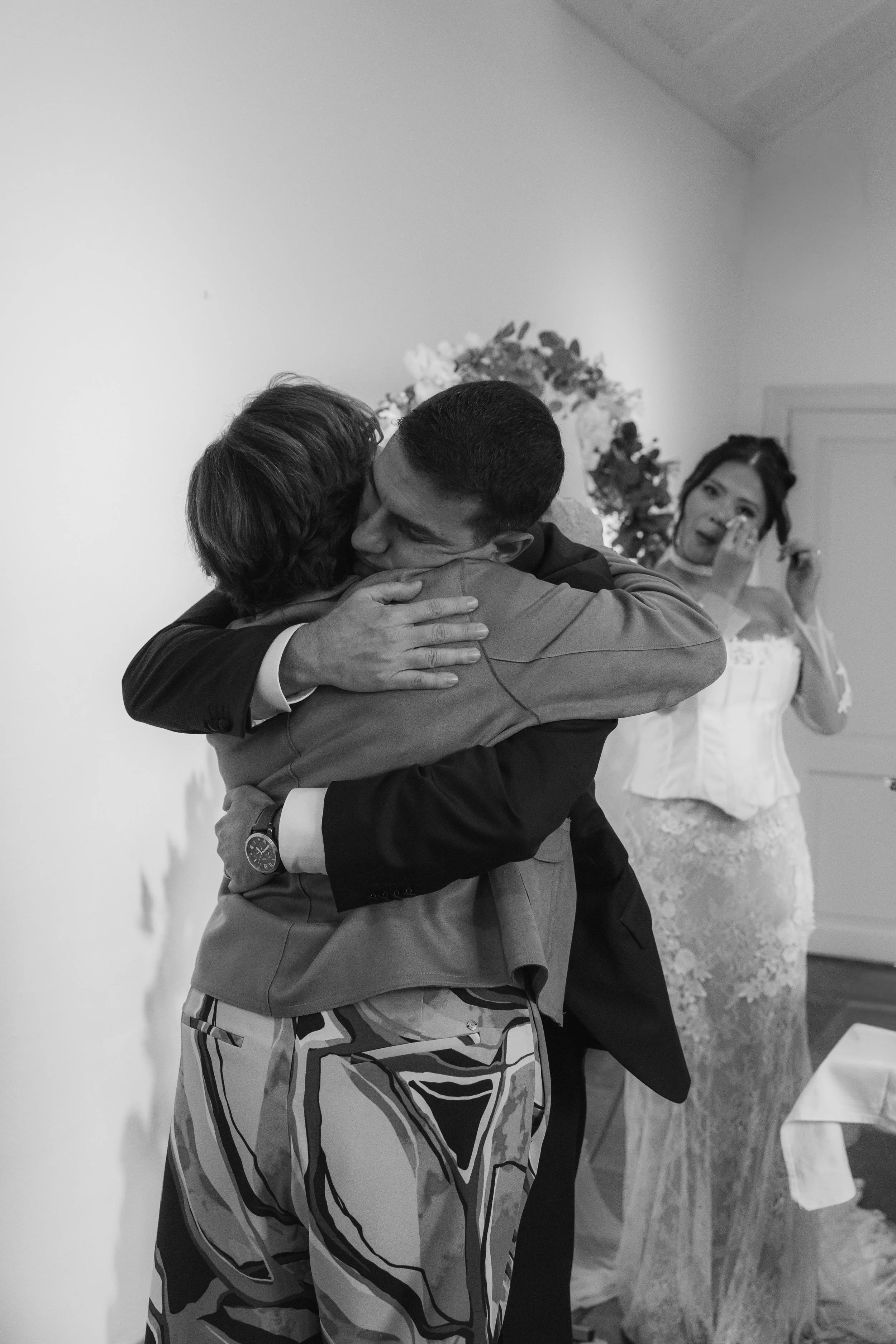 Two men hugging each other, one wearing a suit and patterned pants, the other in a uniform or casual jacket, in a room with a woman in a wedding dress wiping tears in the background.
