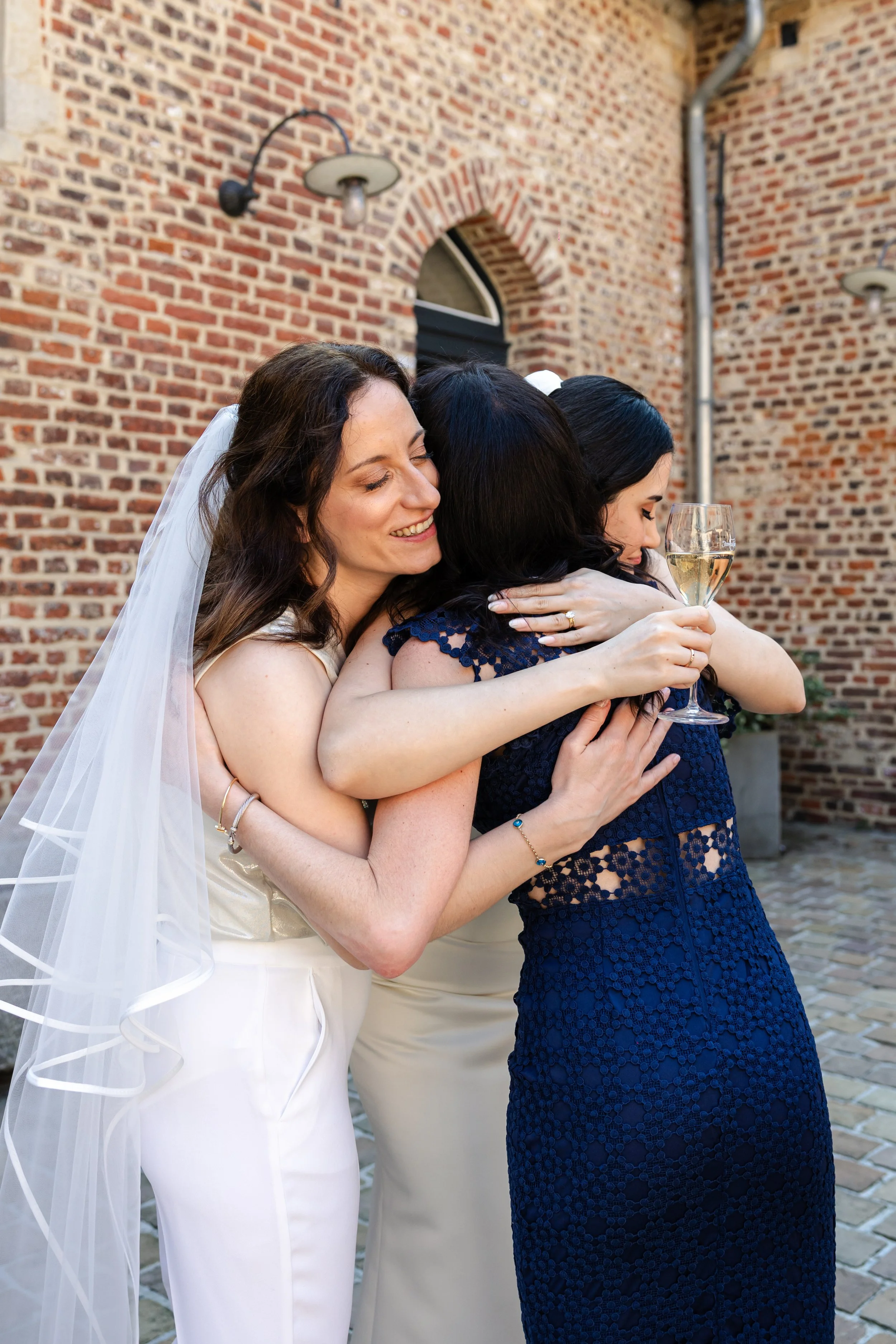 Women hugging at wedding celebration, one woman in wedding dress with veil, others in formal dresses, one holding a glass of champagne, brick building background.