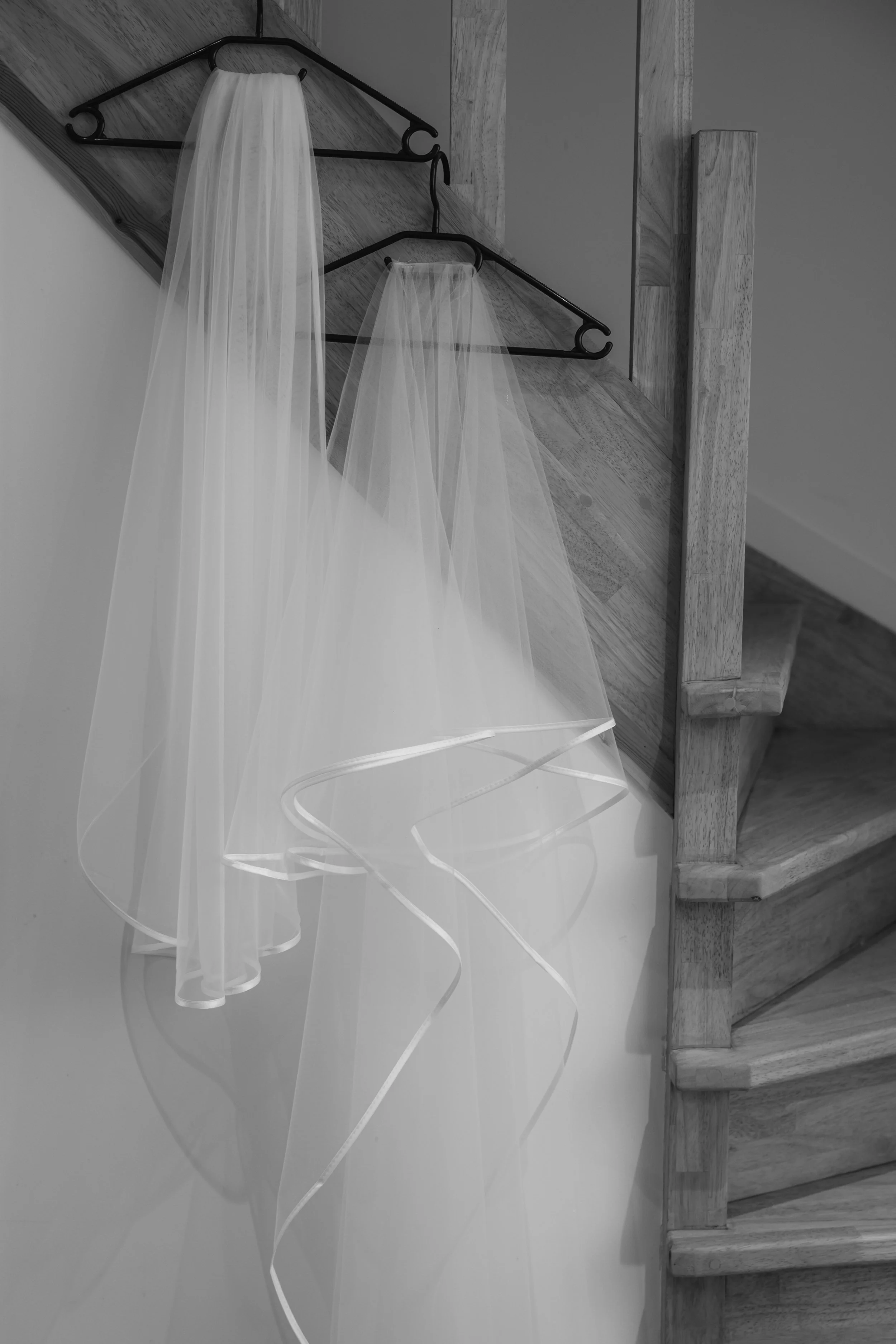 Two white bridal veils hanging on black hangers on a warped wooden wall near a staircase.