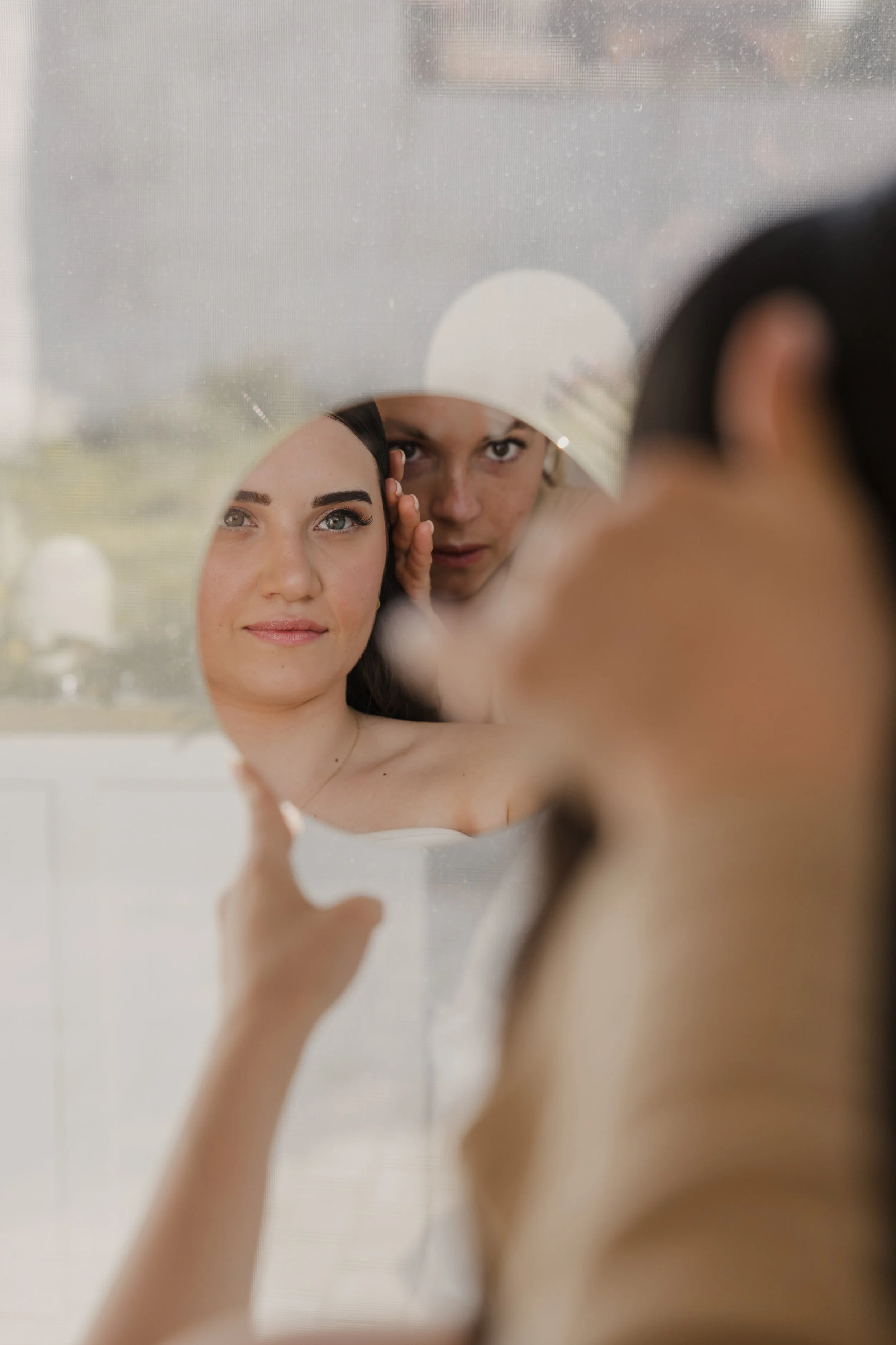A woman is looking at her reflection in a mirror, touching her face, with a makeup artist working on her and partially visible in the background.