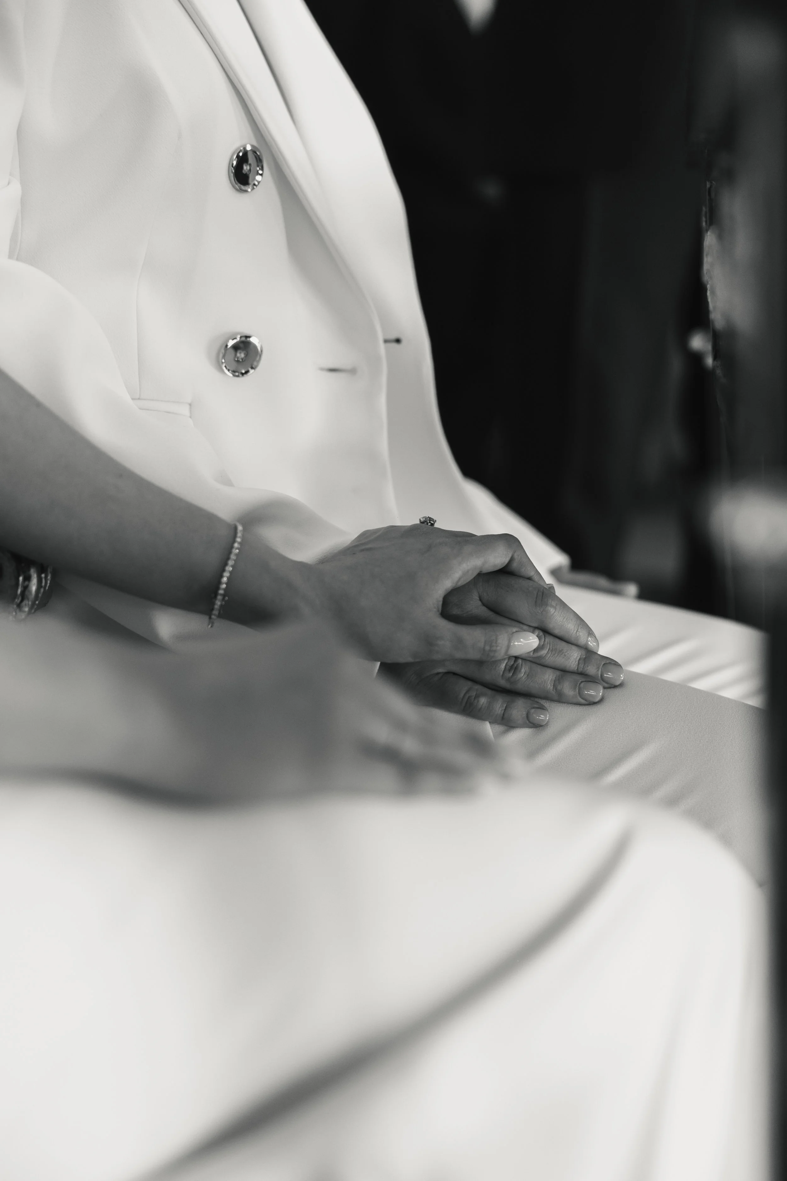 A woman dressed in white sitting with her hands resting on its lap, wearing jewelry. The image is in black and white.