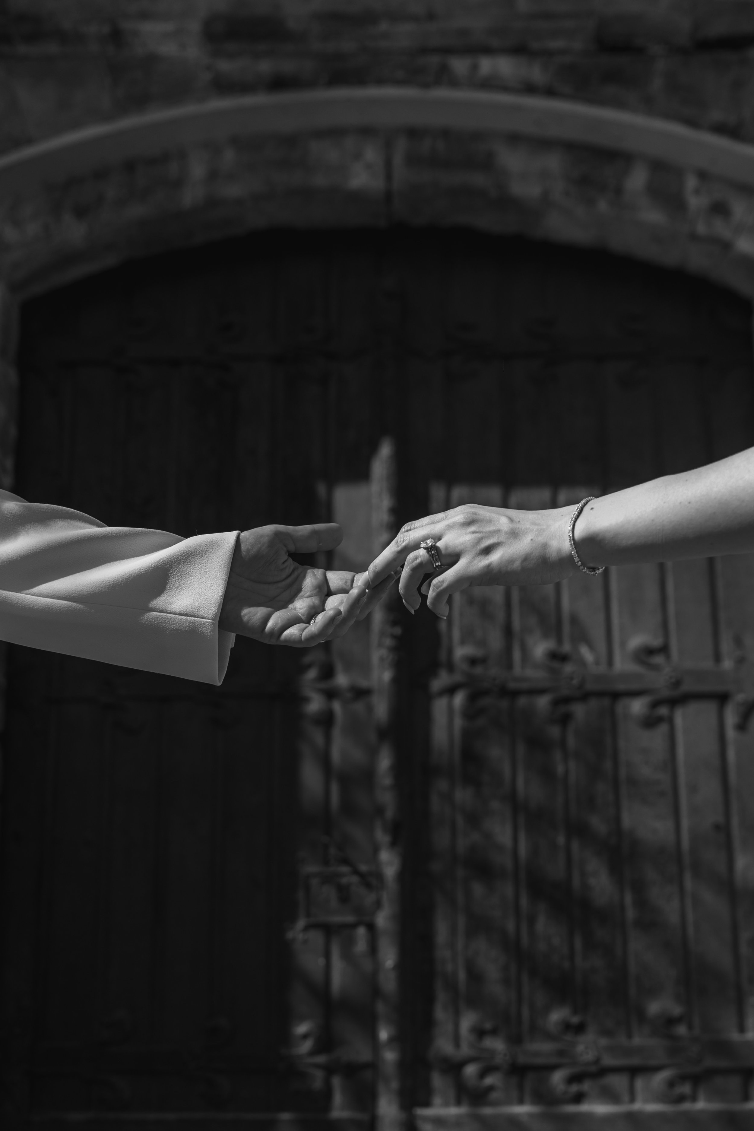 Black and white photo of two hands reaching toward each other, with one hand wearing a ring and bracelet, in front of a large wooden gate.