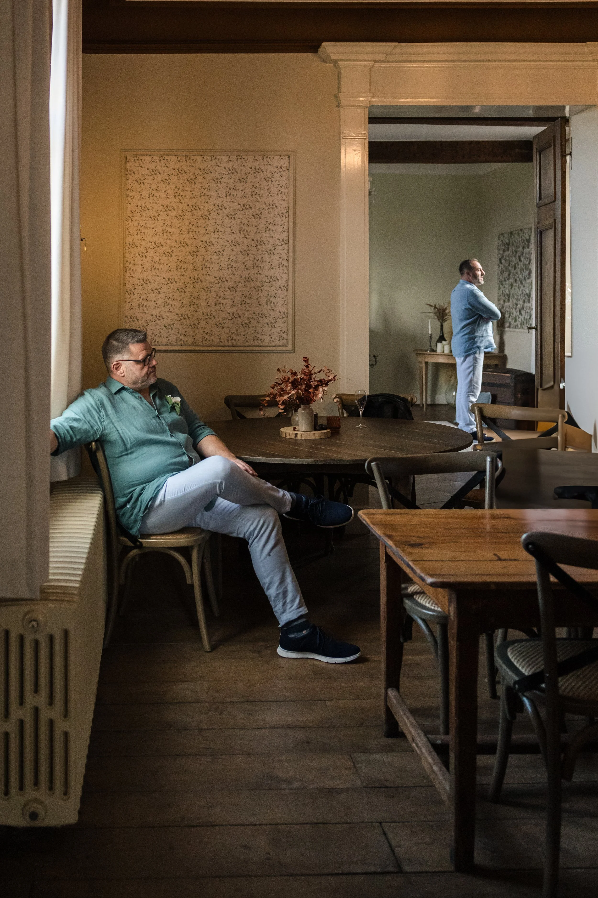 Two men are in a room, separated by a wall and an open doorway. One man with glasses and a teal shirt is sitting against a window on the left, and the other man in a blue shirt is standing in an adjacent room, looking away. The room has wooden floors