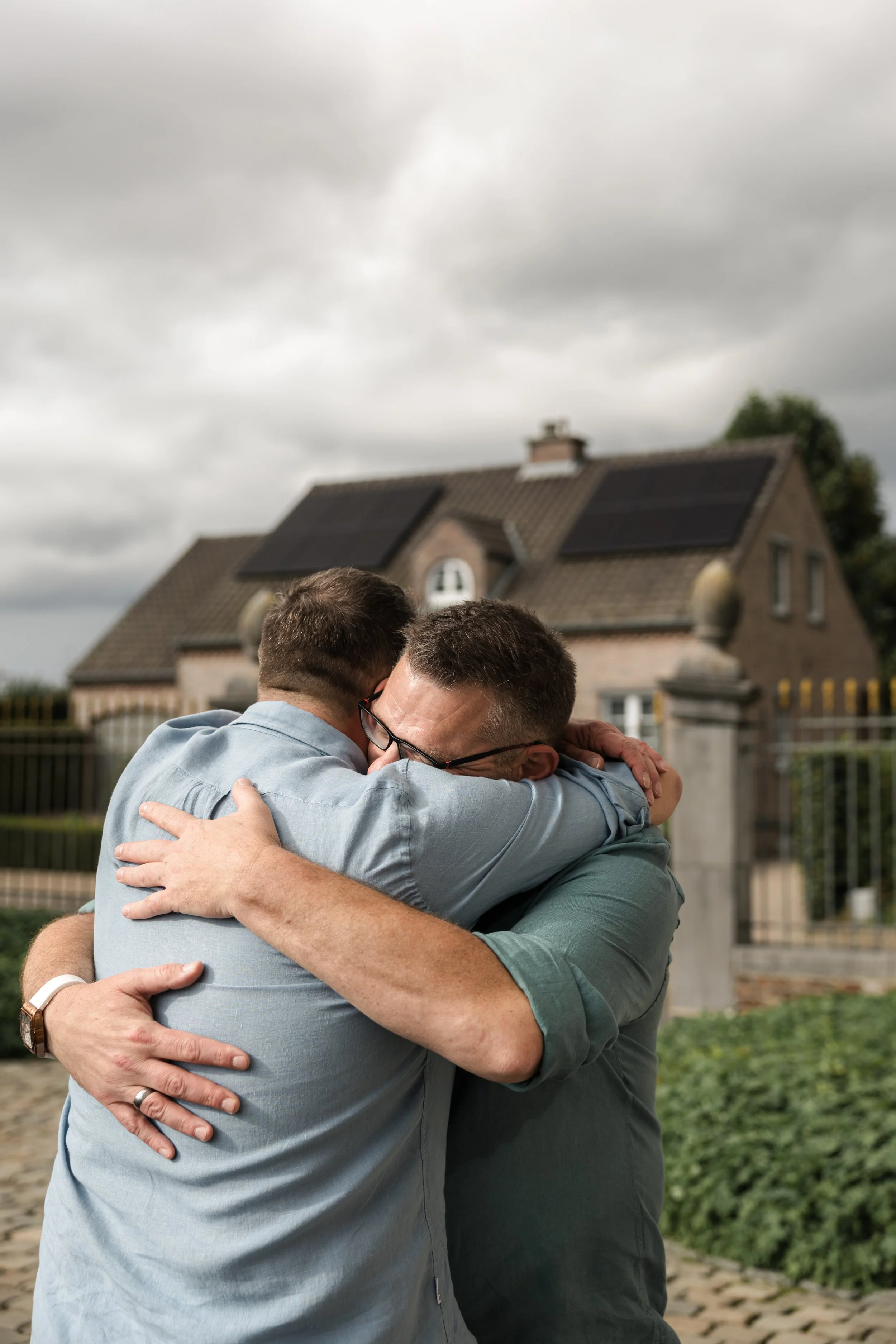 Two men embrace in a hug outdoors, with a house and cloudy sky in the background.