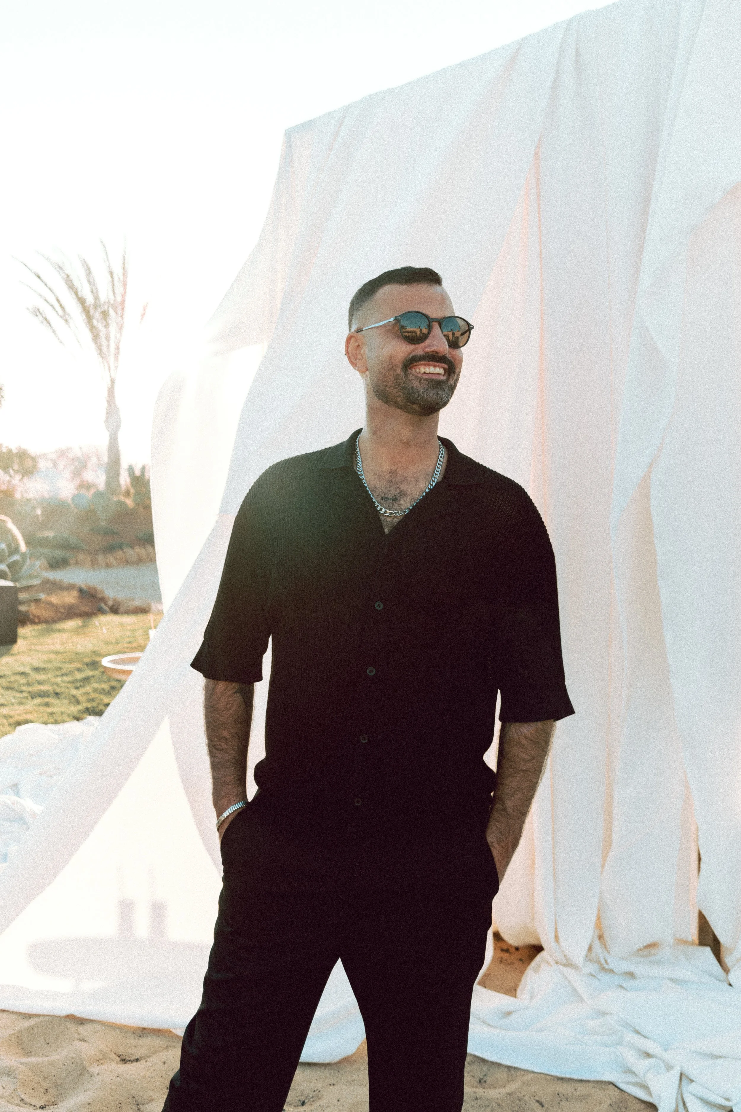 A smiling man with a beard, wearing sunglasses, a black shirt, a silver chain necklace, and a bracelet, standing outdoors near a white fabric backdrop on a sandy beach at sunset.