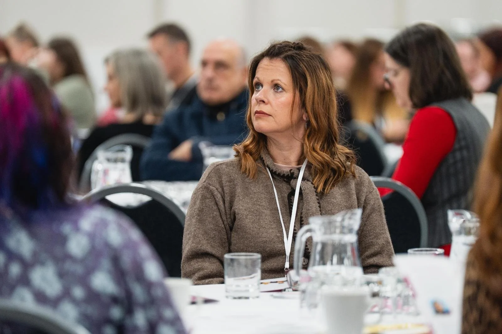A woman with shoulder-length brown hair, wearing a brown sweater and an identification badge, is sitting at a conference table with a glass pitcher and glassware in front of her. She appears attentive, attending a conference or meeting with other div