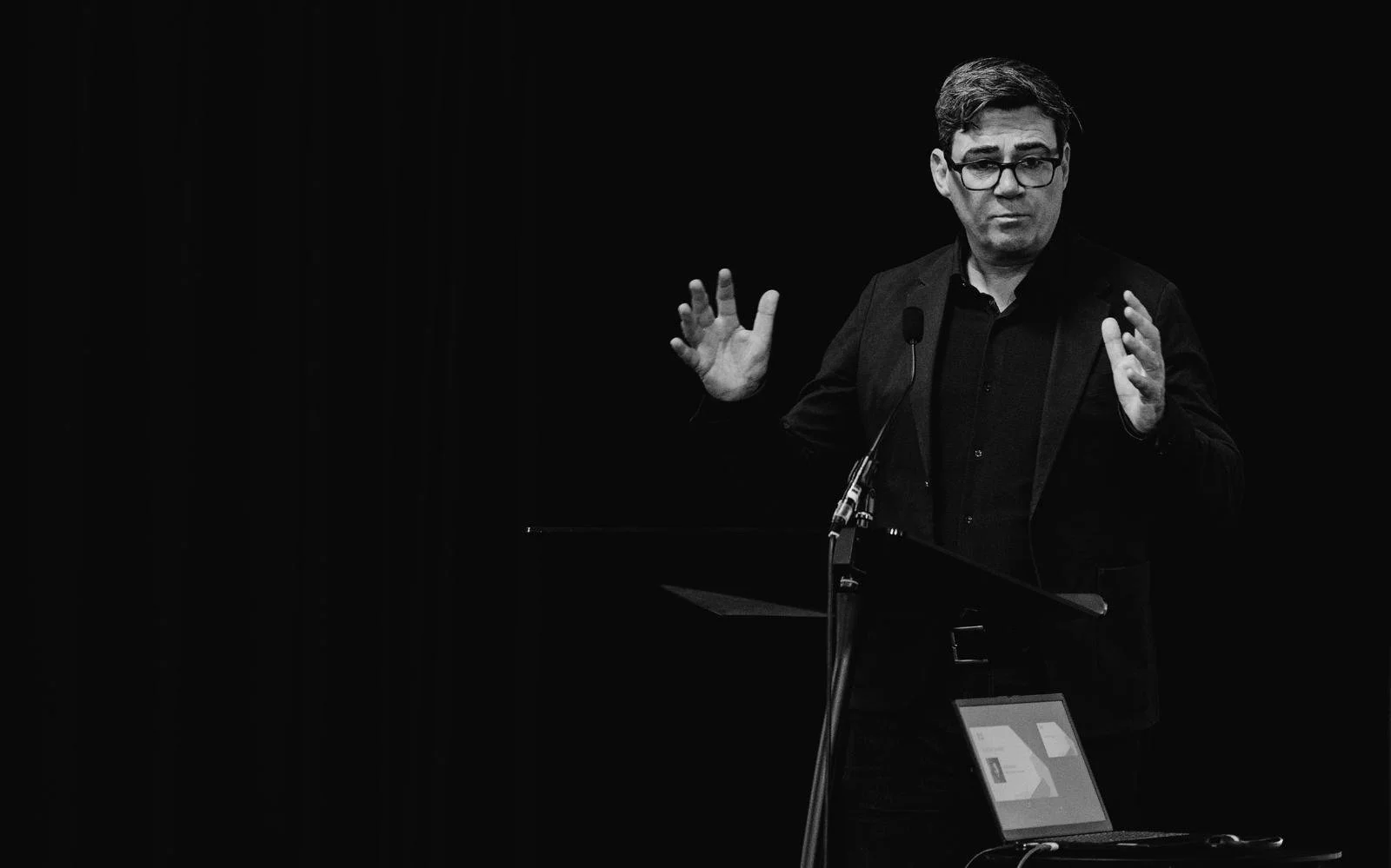 A man wearing glasses and a dark suit stands behind a podium, gesturing with his hands during a presentation or speech against a dark background.