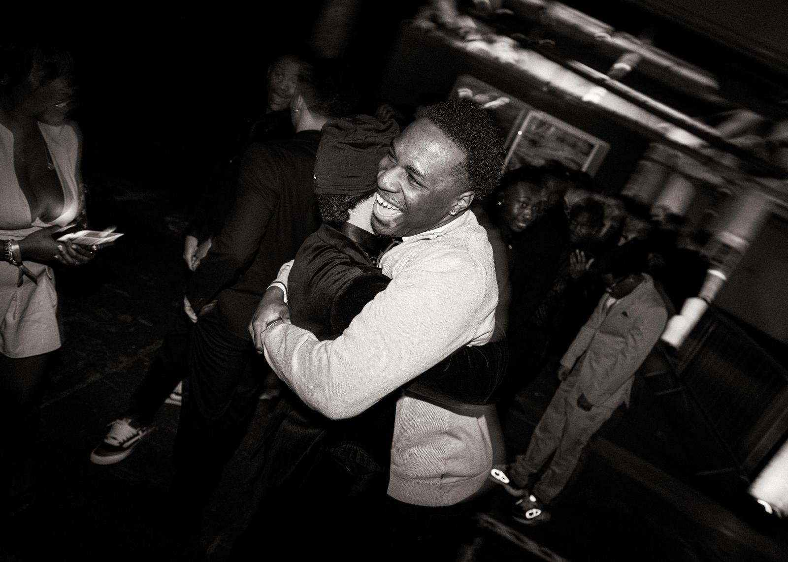 Two men embracing and smiling at a lively party, surrounded by other people, in a dark indoor setting.