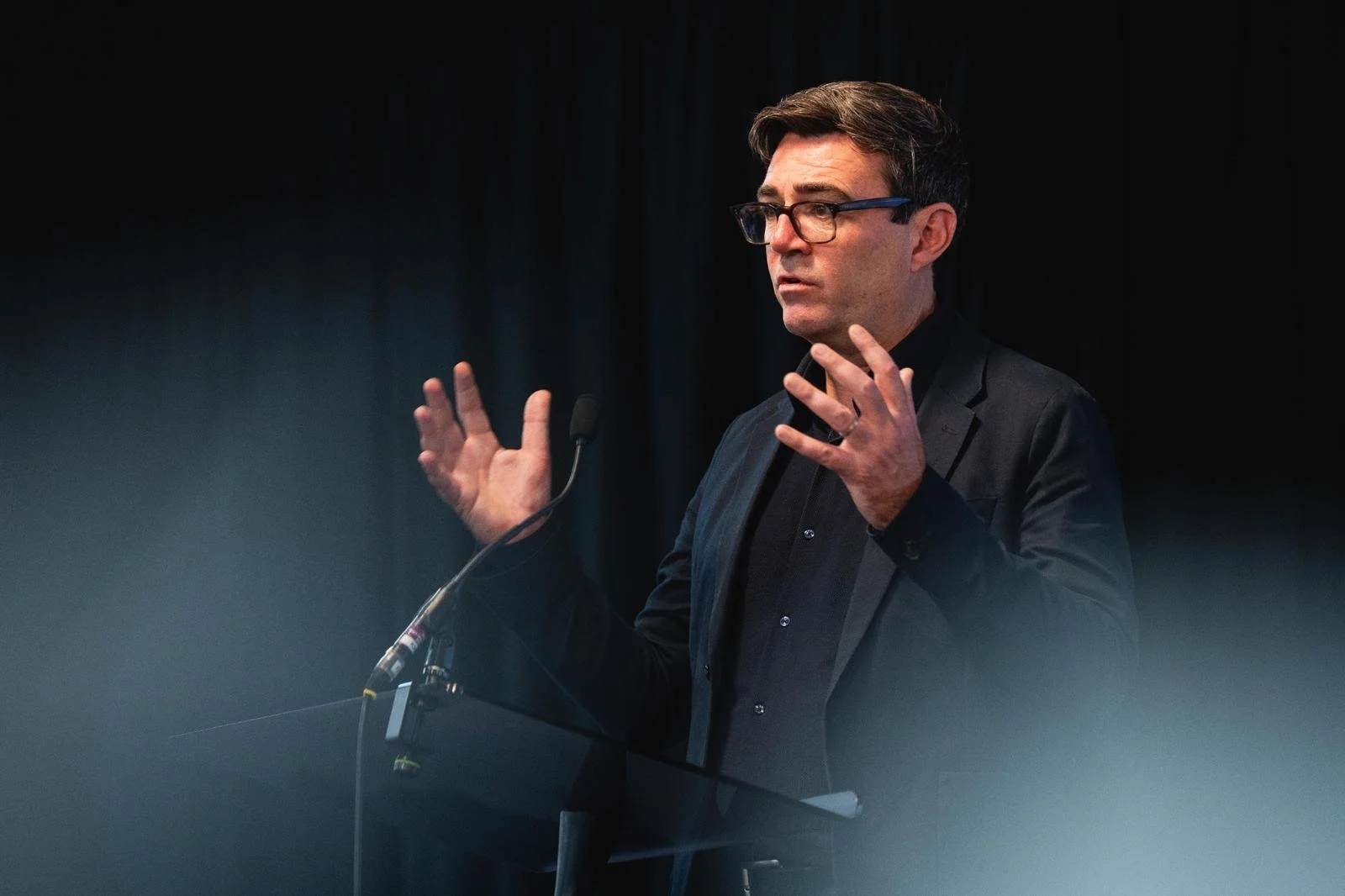 A man wearing glasses and a dark blazer speaking at a podium with a microphone, gesturing with his hands.