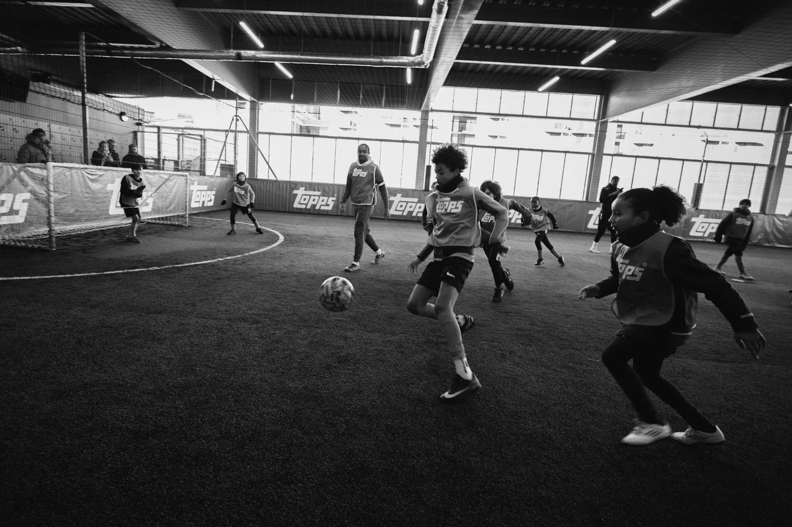 Children playing indoor soccer on a turf field with coaches and spectators nearby, large windows in the background letting in natural light.