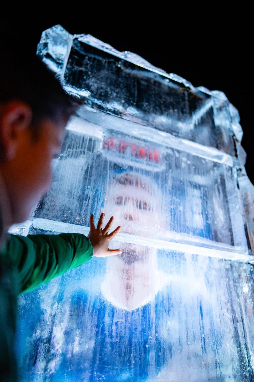 Child reaching out to touch a large block of ice, with the child's reflection visible in the ice.