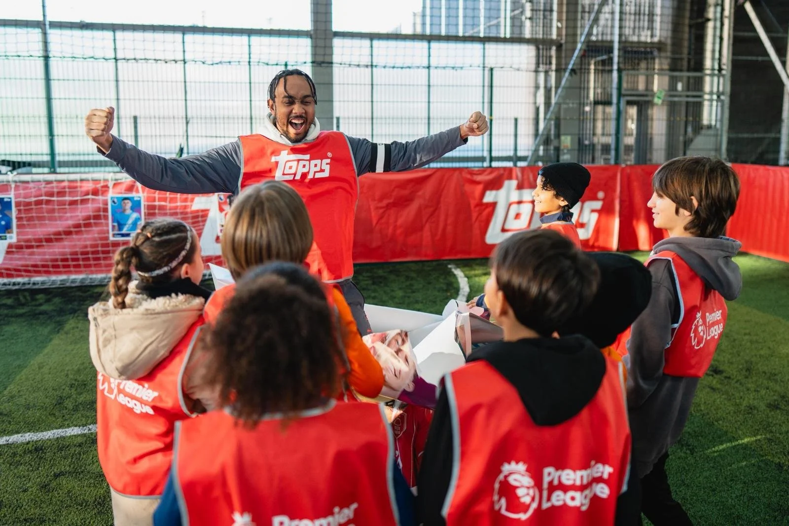 A soccer coach or instructor with long braids, wearing a red vest with a 'Topps' logo, is celebrating with children during a soccer training session inside an indoor facility. The children are wearing red vests with 'Premier League' logos and are sit