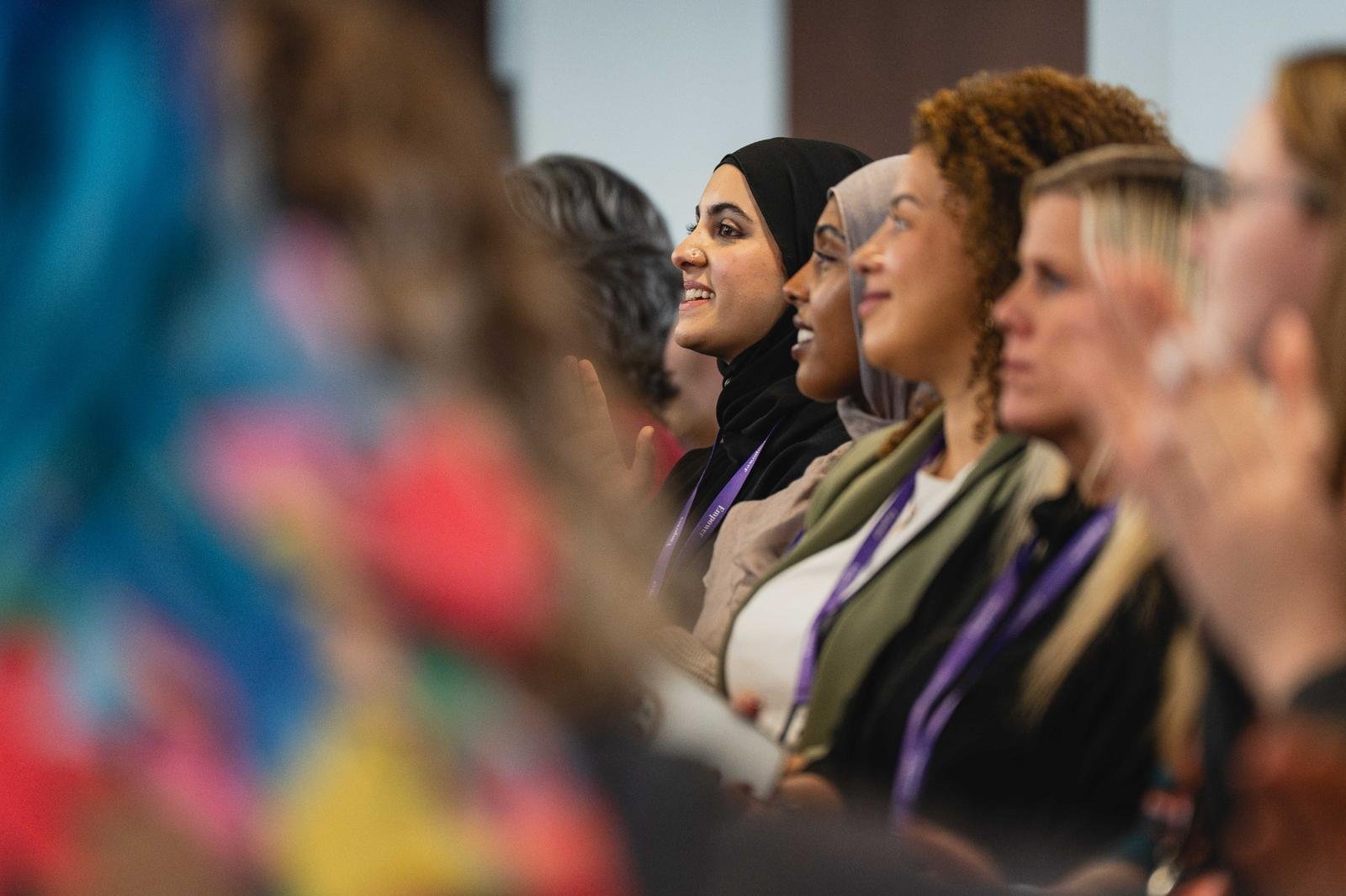 A diverse group of women sitting side by side, attending a conference or seminar, listening attentively with slight smiles.