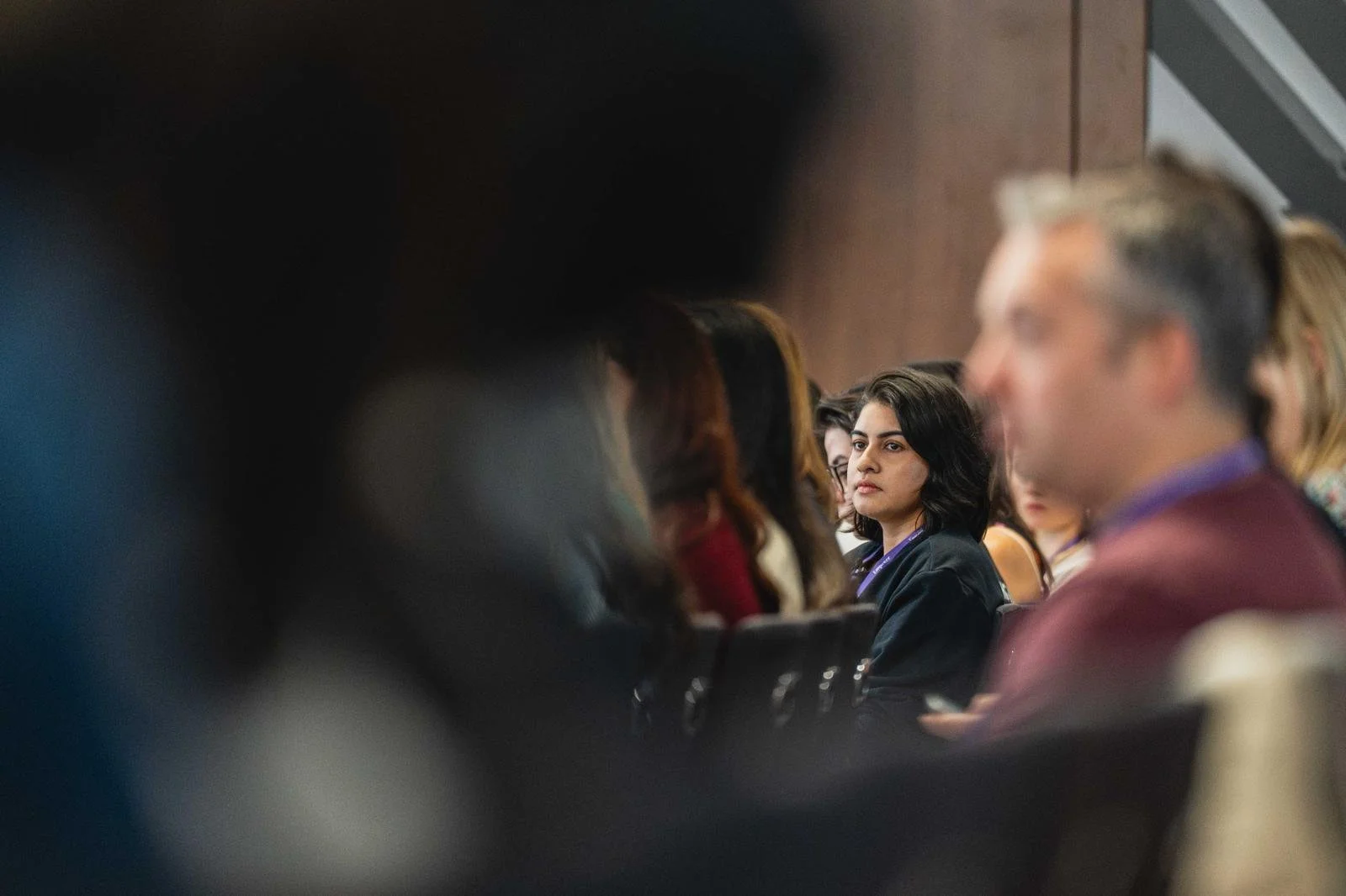 A woman with dark hair and a serious expression sitting among a blurred crowd in a conference or meeting room.
