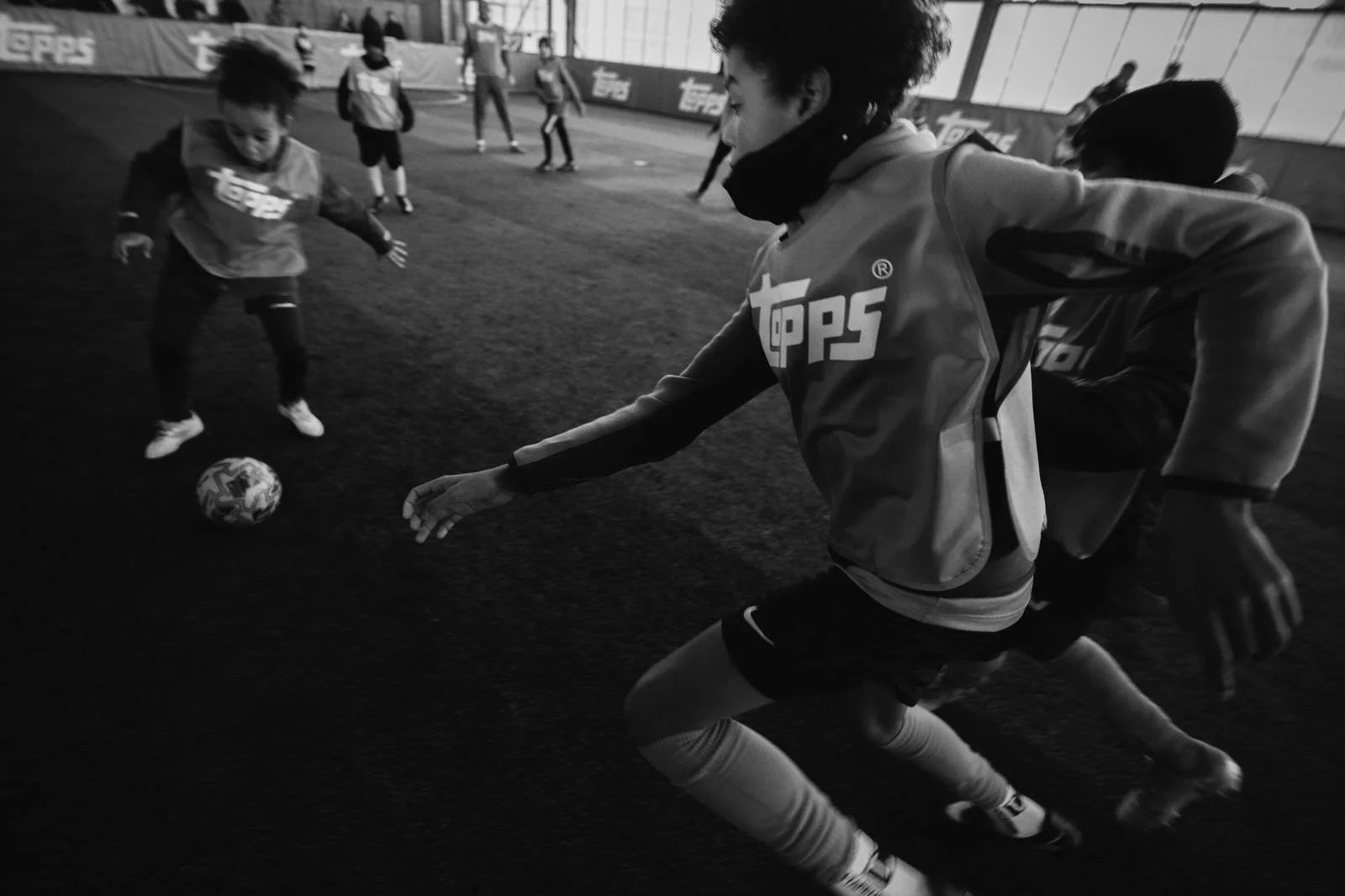A young soccer player in motion on an indoor field, wearing a jacket with a logo, as other children and coaches play in the background.
