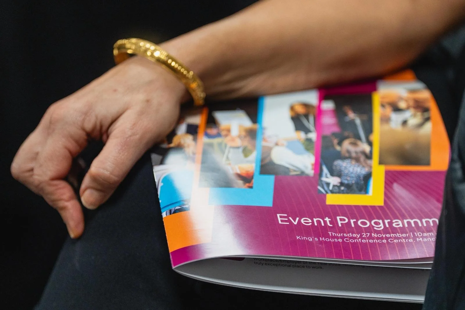 A hand with a gold bracelet holding a colorful event program flyer with photos, text, and a pink background.