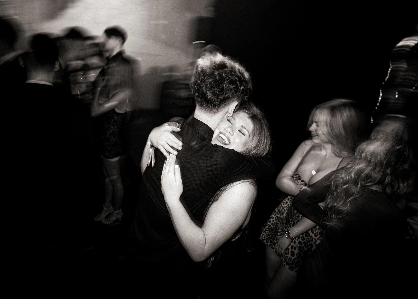 A couple hugging tightly and smiling at a social event with other people in the background.
