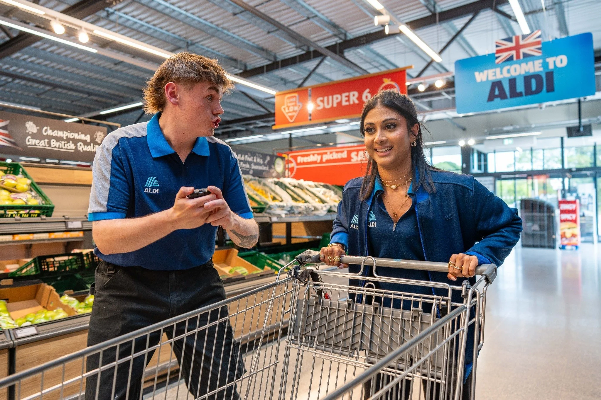 A male and female cashier, both wearing blue ALDI uniforms, engaging with each other at a supermarket checkout counter. The male cashier is showing something on his phone, and the female cashier is looking at him, smiling. There is a shopping cart in