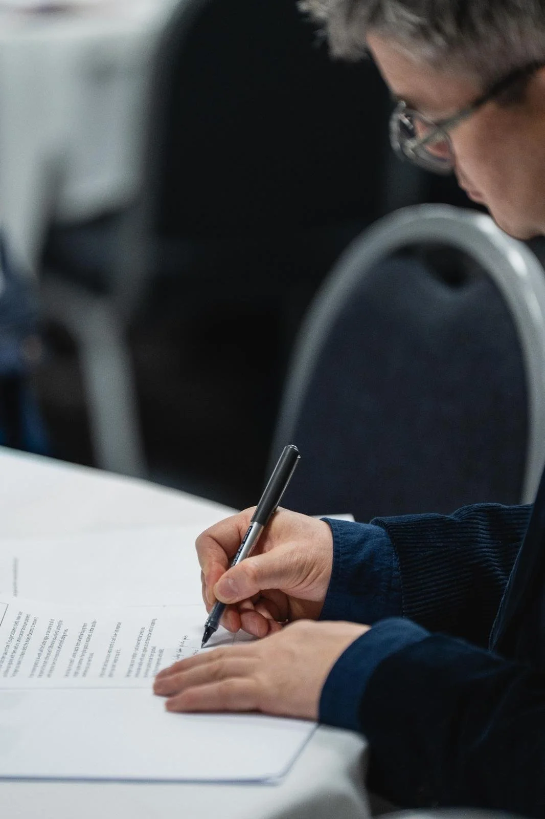 A person with glasses and a dark blue sweater is sitting at a table, writing on a document with a black pen. The background shows some blurred chairs and tables.