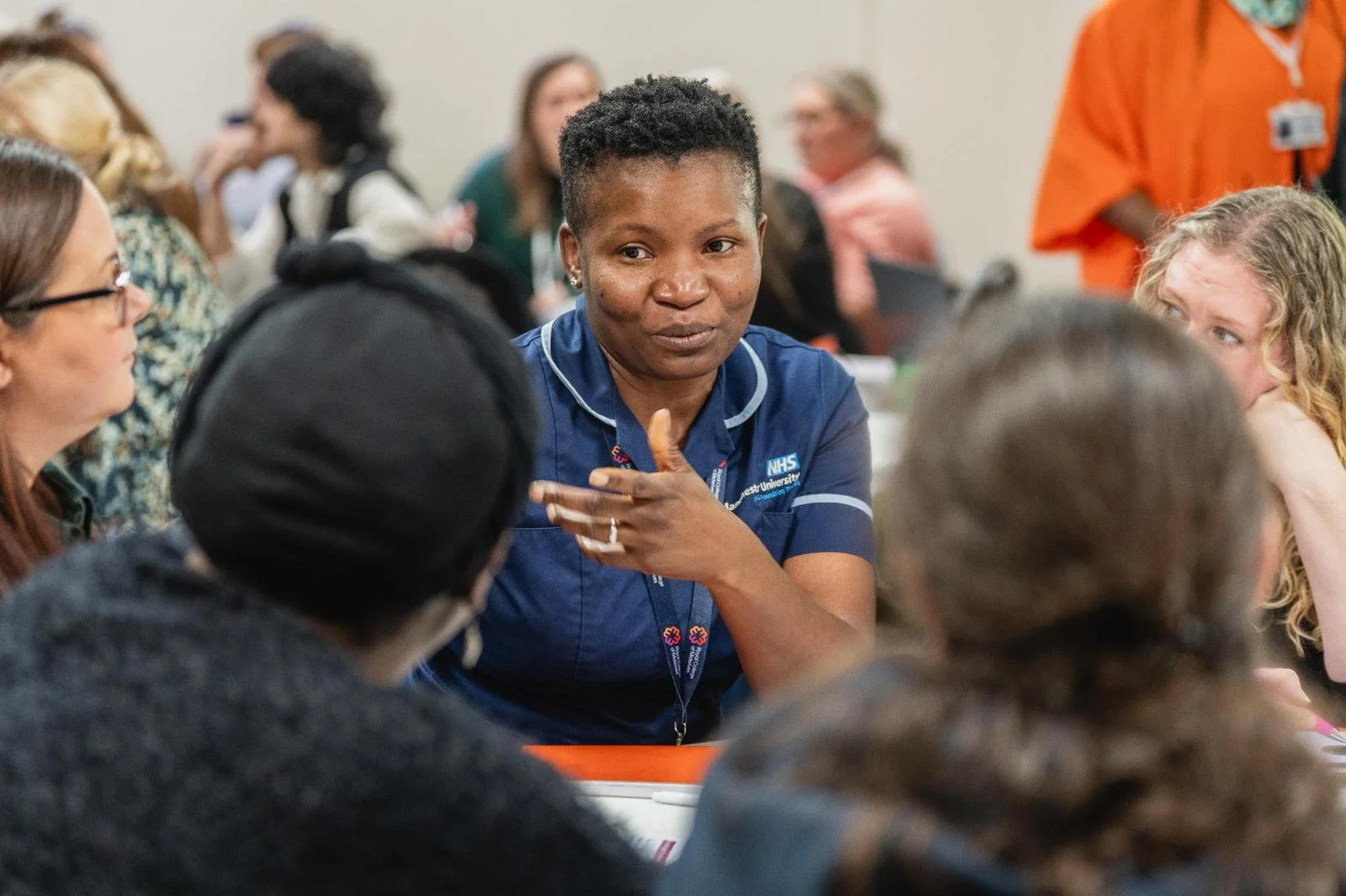 A woman in a NHS nurse uniform speaking and gesturing during a discussion at a roundtable with diverse participants at a conference.