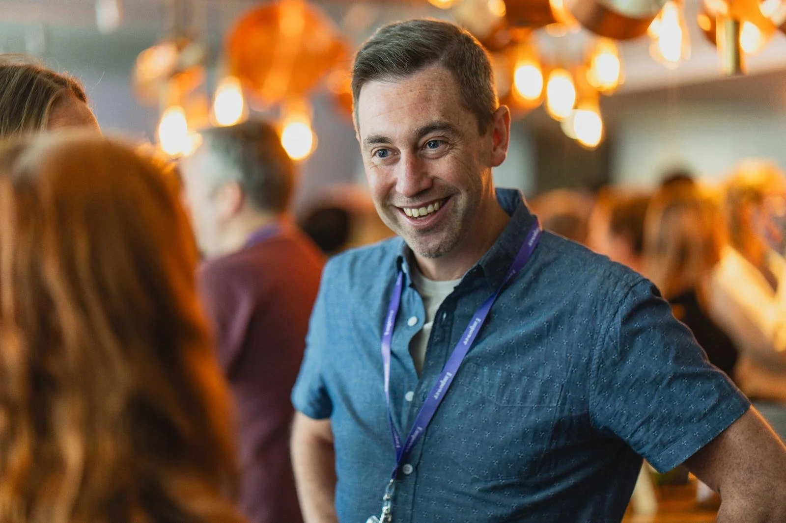 A man smiling at a social event with warm lighting and people in the background.