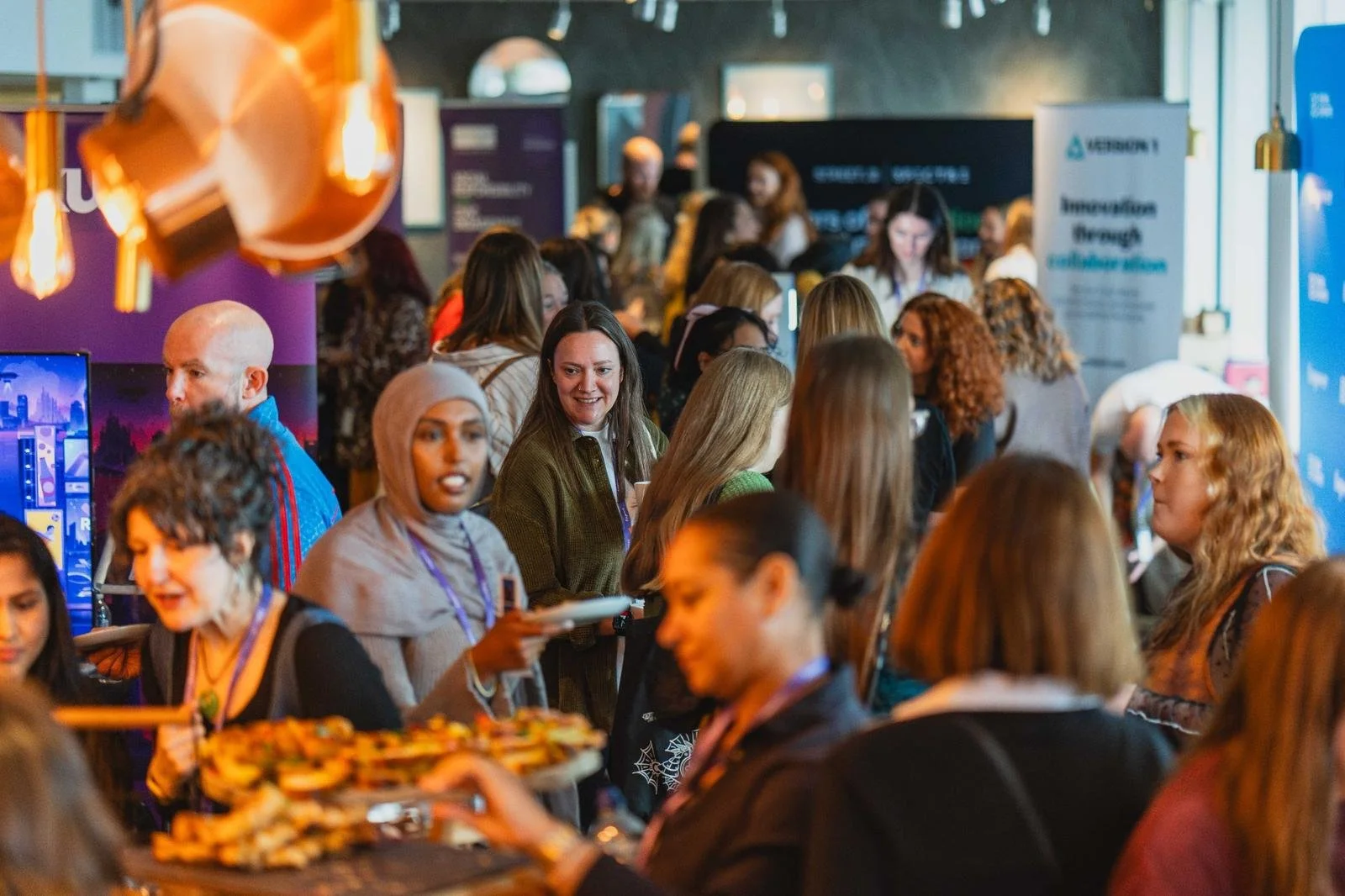 Crowd of people at a crowded indoor event with displays and banners, some engaging in conversation and others serving food.