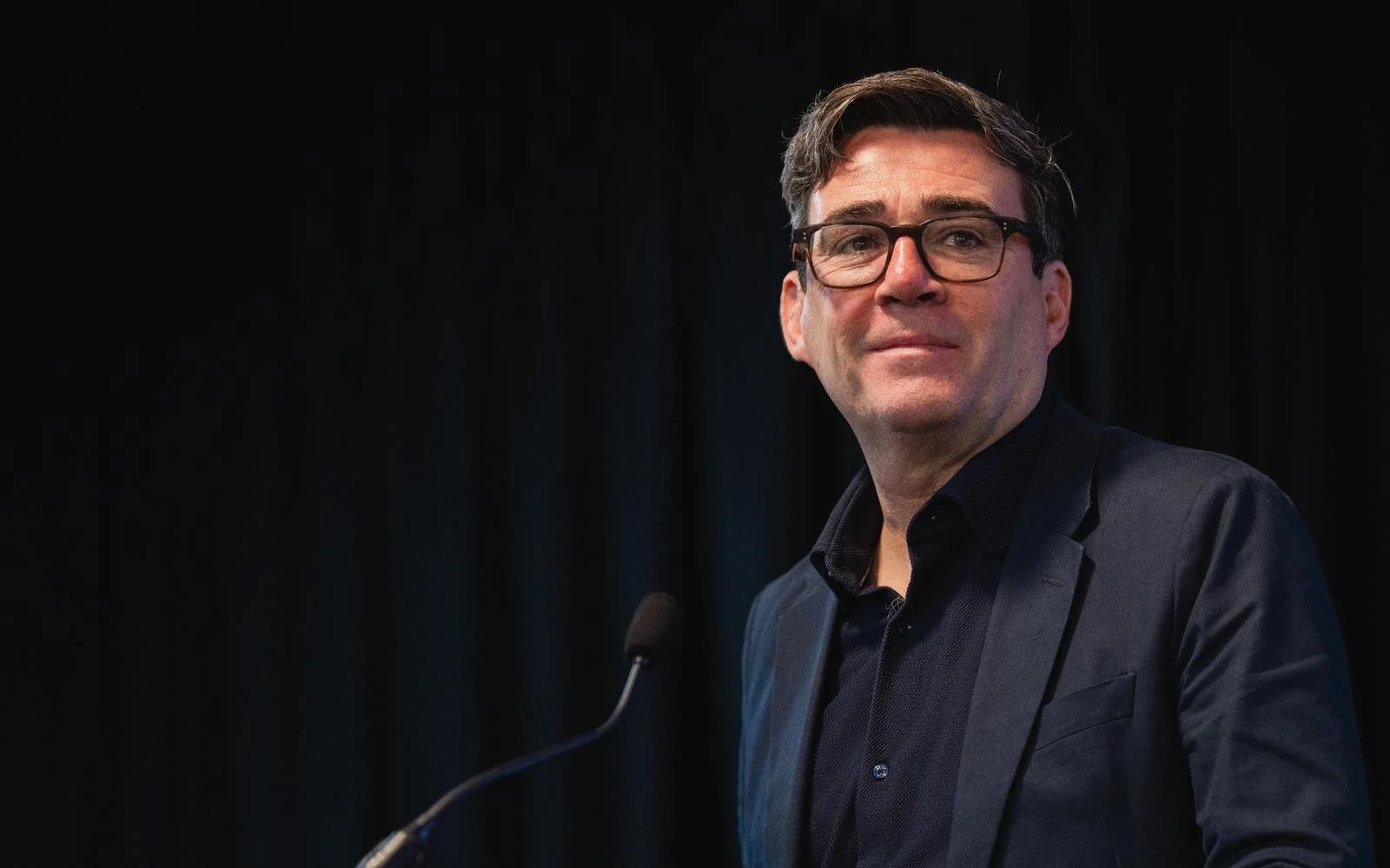 A man with glasses and dark hair wearing a dark suit and shirt, standing at a microphone against a dark background.