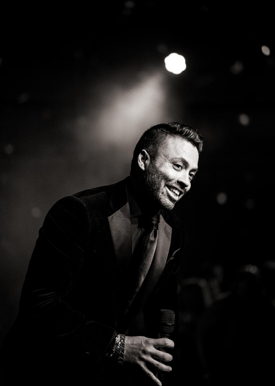 A man holding a microphone on stage, smiling and leaning forward, wearing a dark suit and tie, with a spotlight behind him.