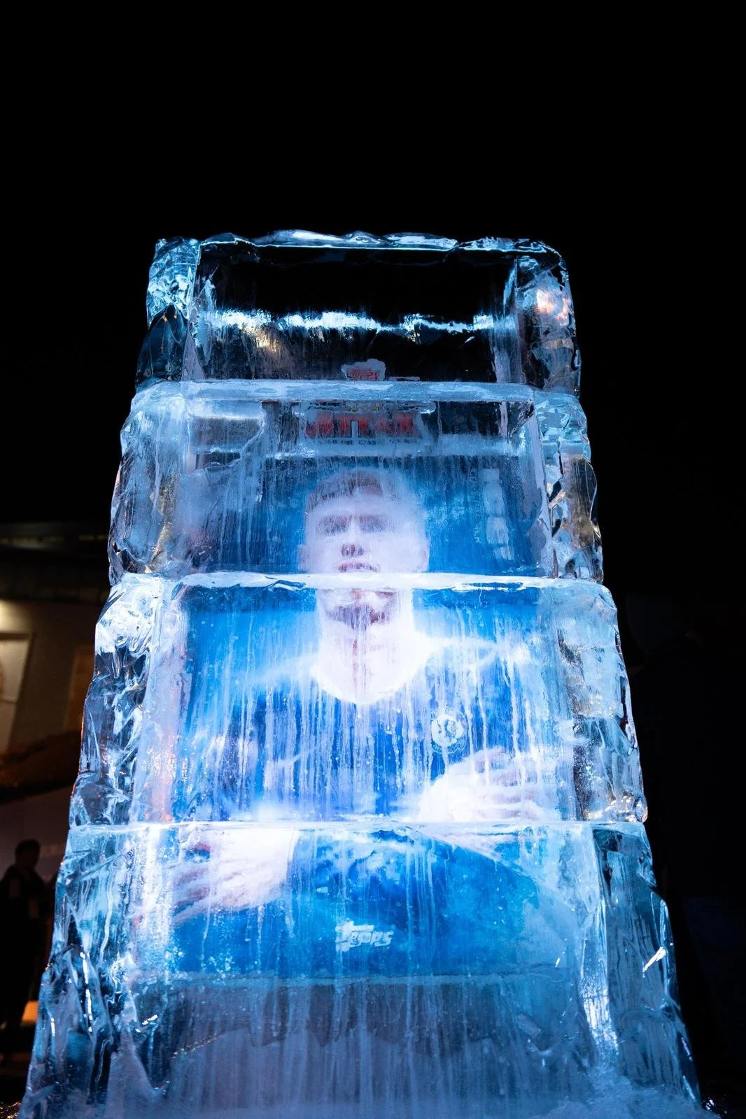 A person behind a large vertical ice sculpture illuminated with blue light at night.