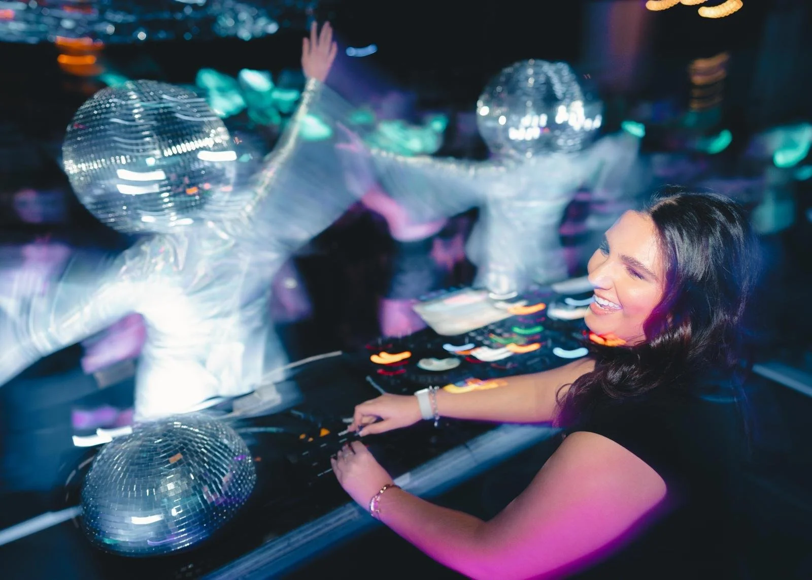 A woman smiling while DJing at a nightclub with disco balls hanging from the ceiling, colorful lights reflecting on her face and the DJ equipment.