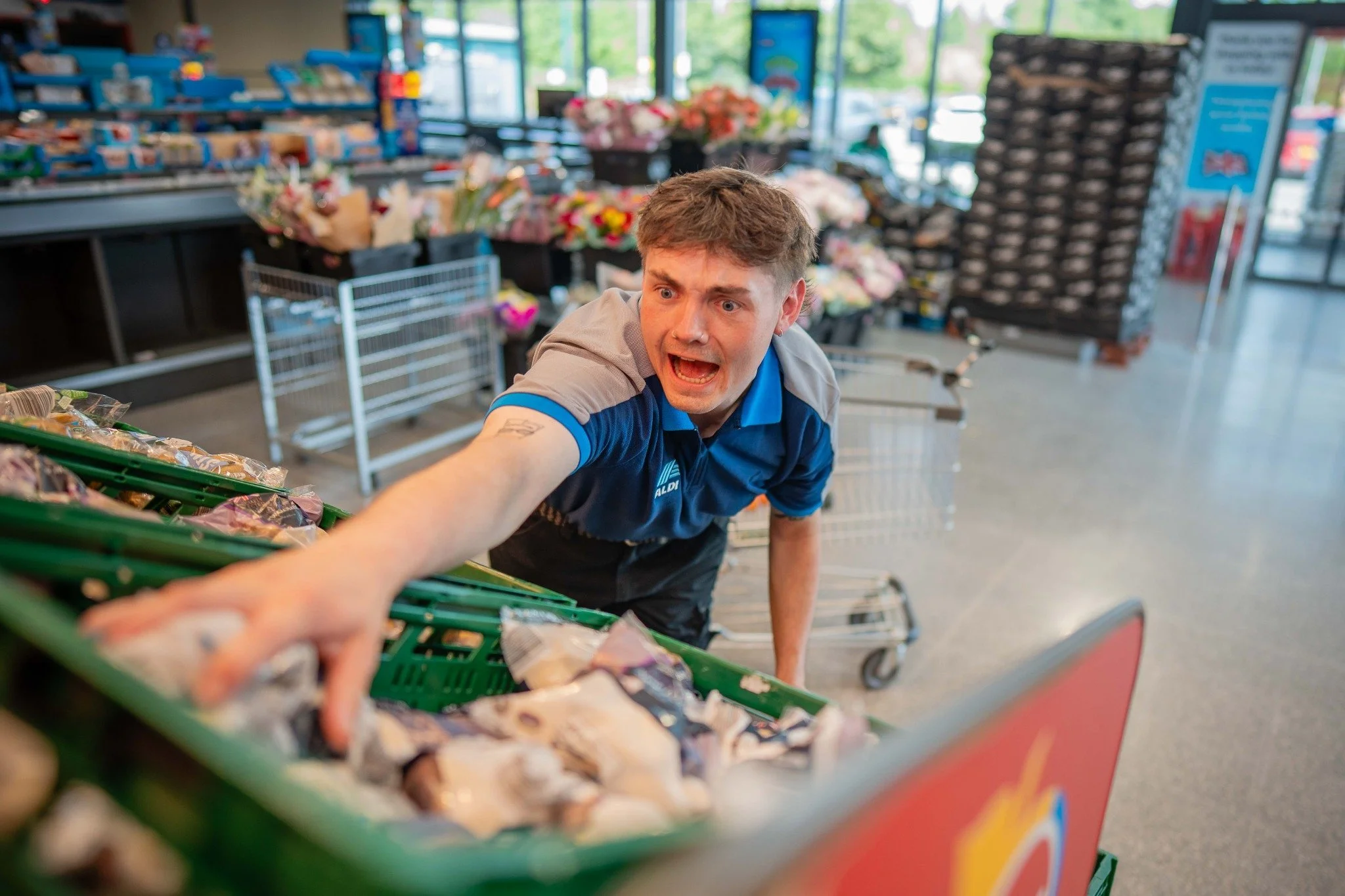A man reaching into a green shopping basket filled with snack bags inside a grocery store with flower bouquets and shelves in the background.