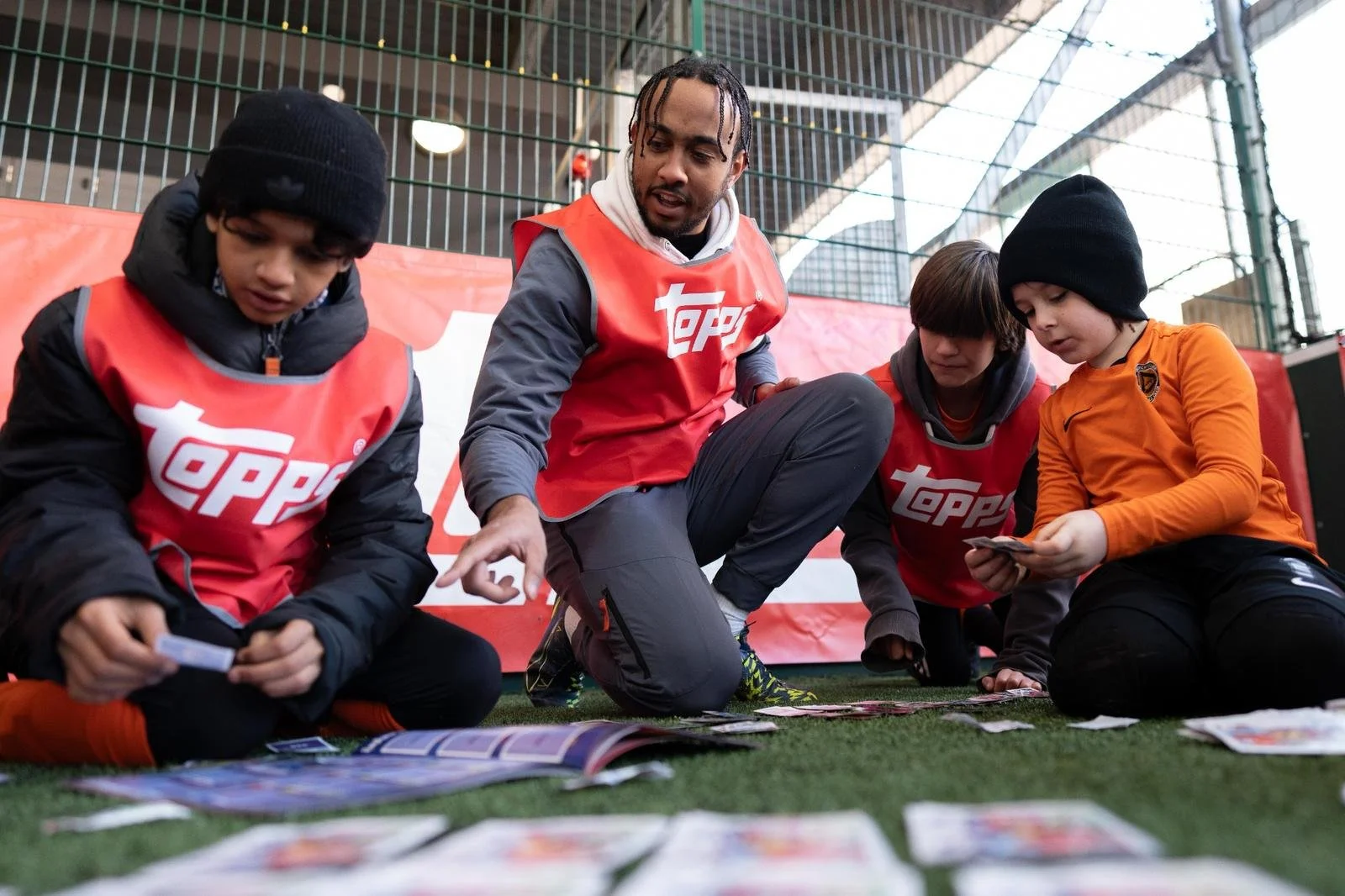 A man with braided hair wearing a red vest with the Topps logo instructs three children, all wearing jackets and beanies, as they look at trading cards spread on the ground. They are on a green turf field near a gated area.
