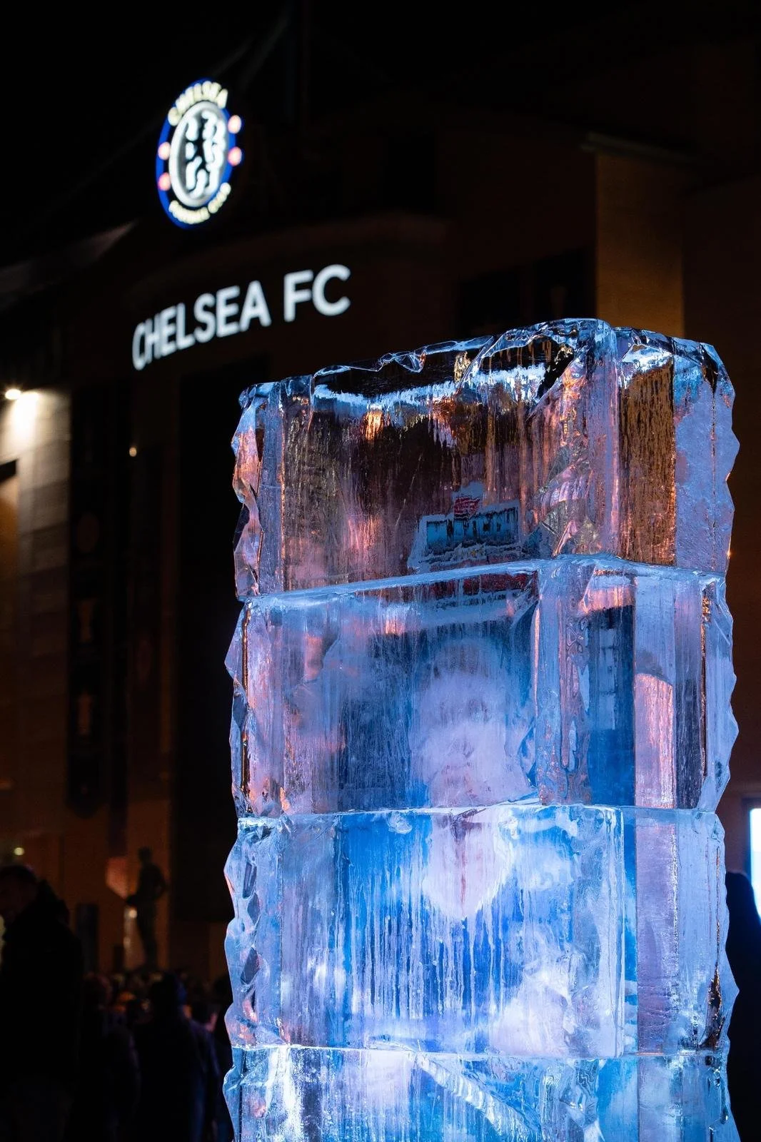 A tall ice sculpture in front of Chelsea FC stadium sign illuminated at night with fans in background.