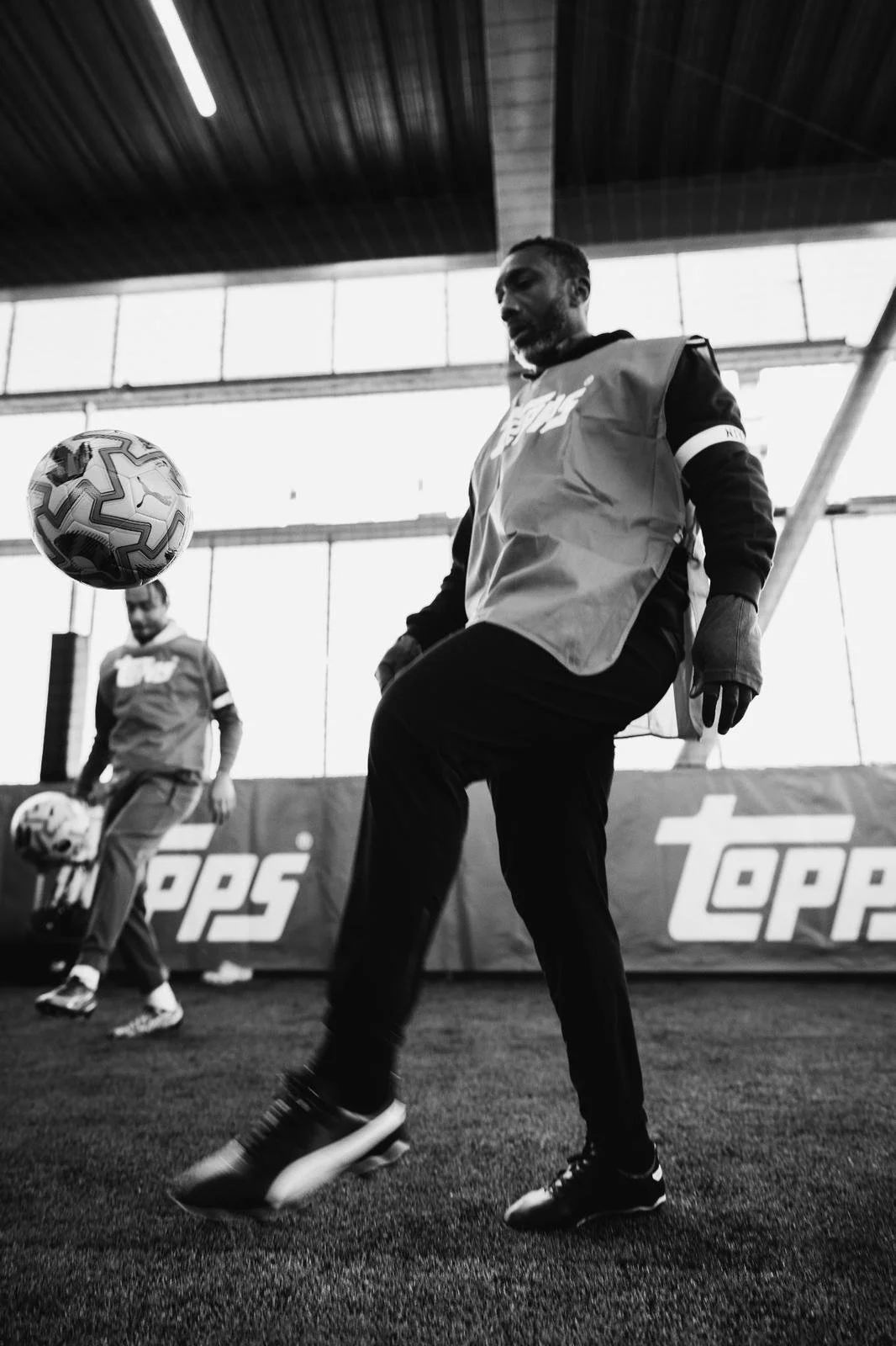 Two men practicing soccer indoors; one is kicking a soccer ball while the other holds a soccer ball and observes.