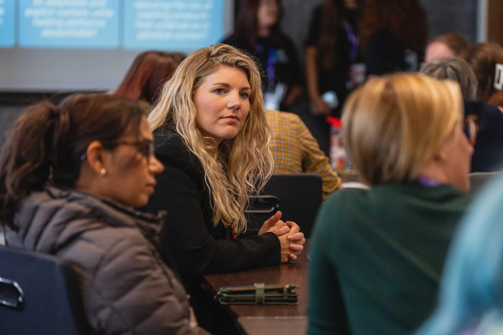 A woman with blonde, wavy hair wearing a black blazer sits at a table during a conference or meeting, surrounded by other women.