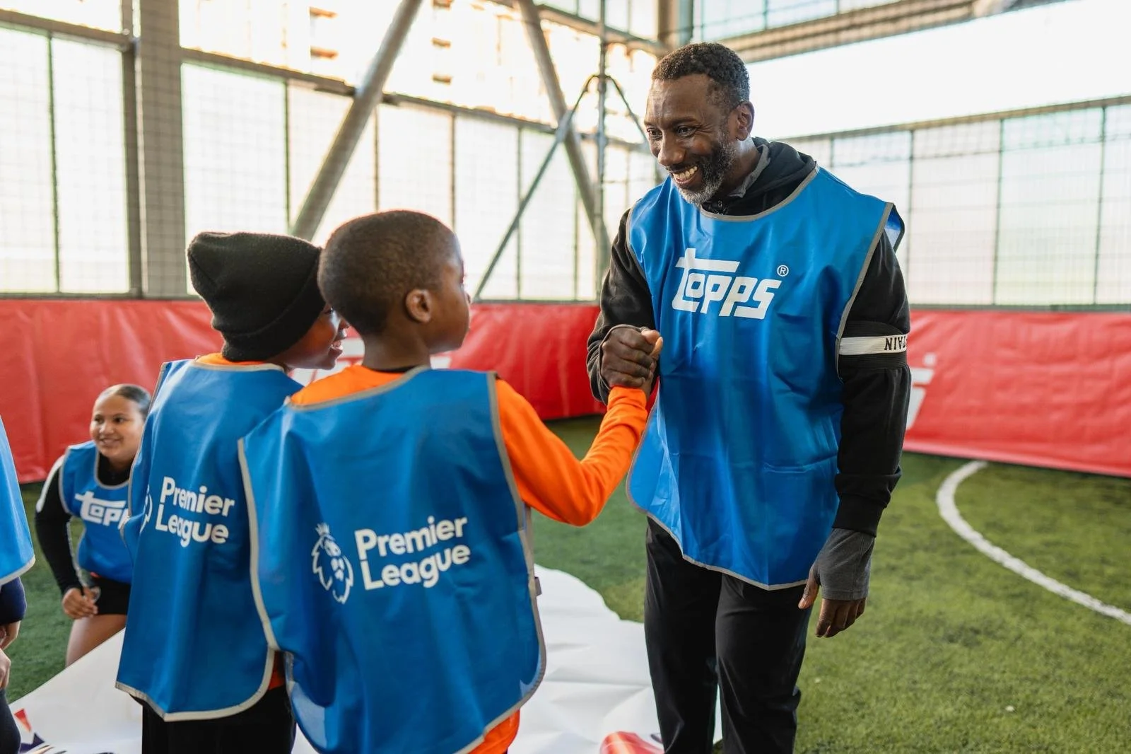 A man in a blue Topps vest is greeting two children on a soccer field. The children are also wearing blue Topps vests that say 'Premier League.' One child is wearing a black beanie and an orange shirt, and the other is wearing an orange shirt. In the