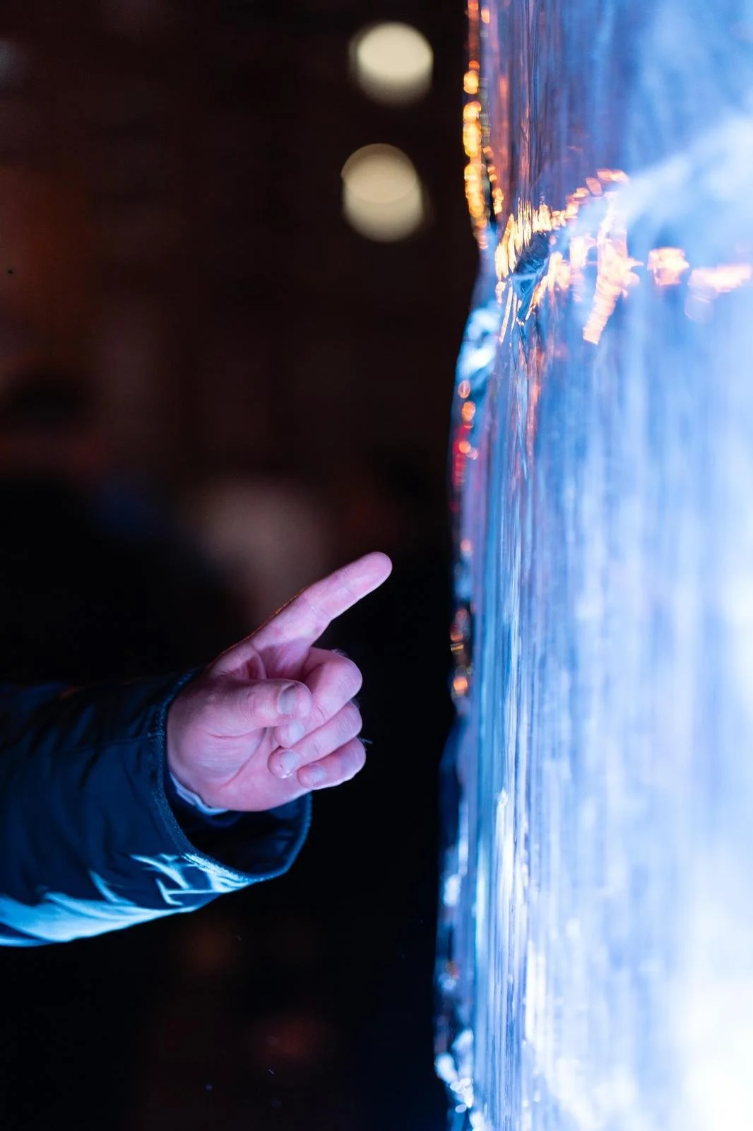 Close-up of a person's hand with a puffy jacket, reaching out to touch a large, curved ice surface with colorful reflections, at night.
