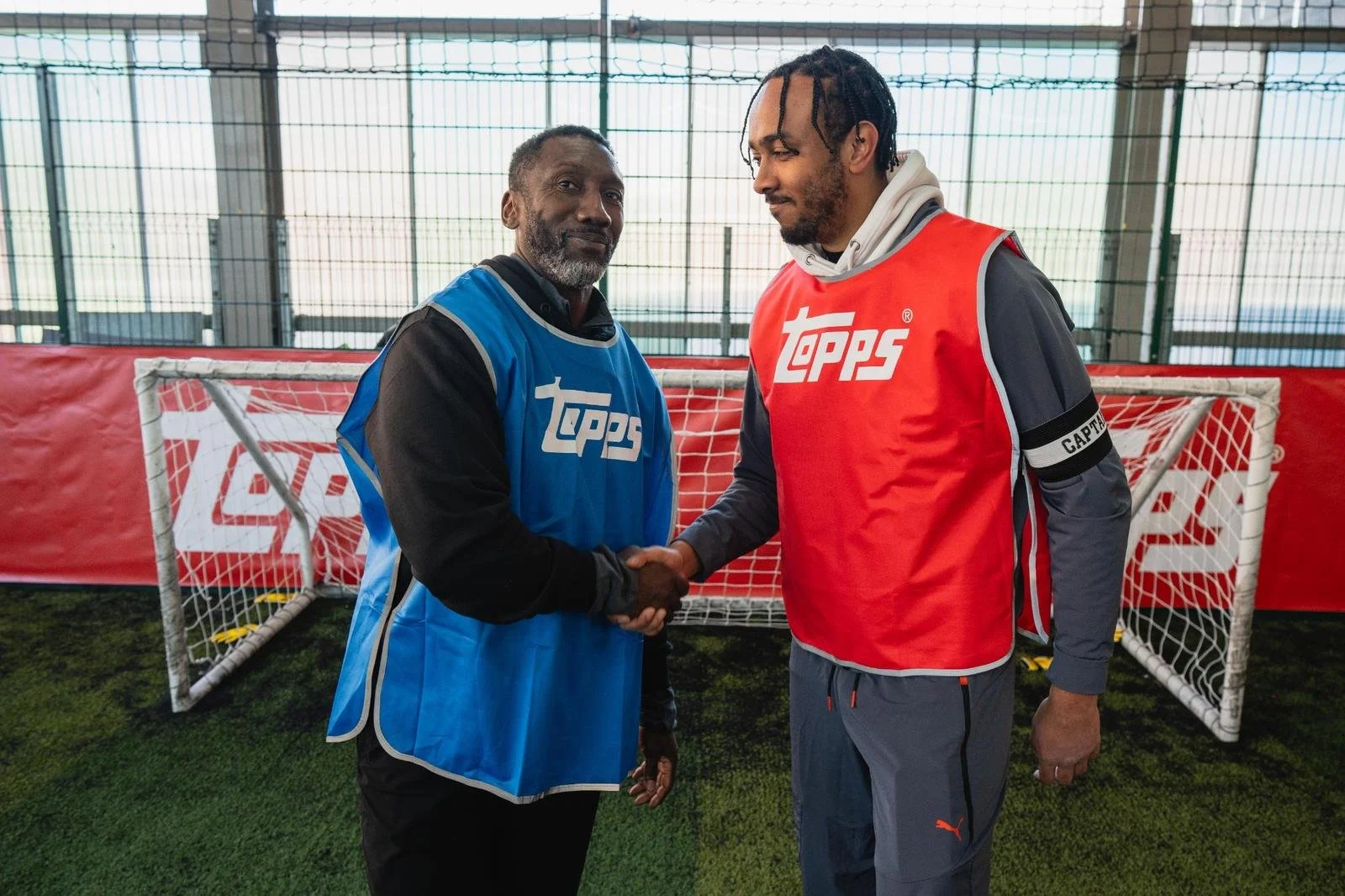 Two men wearing blue and red sports vests shaking hands on an indoor soccer field.