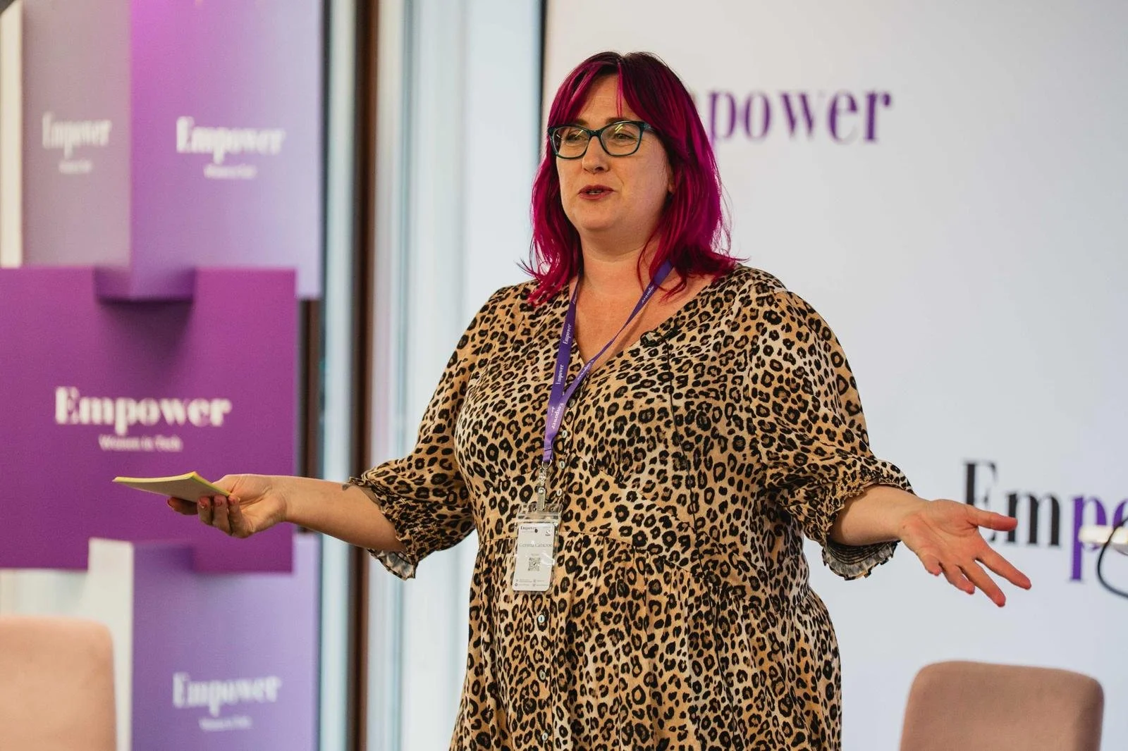 A woman with pink hair, glasses, and a leopard print dress speaking at an event, with purple banners that say 'Empower' in the background.