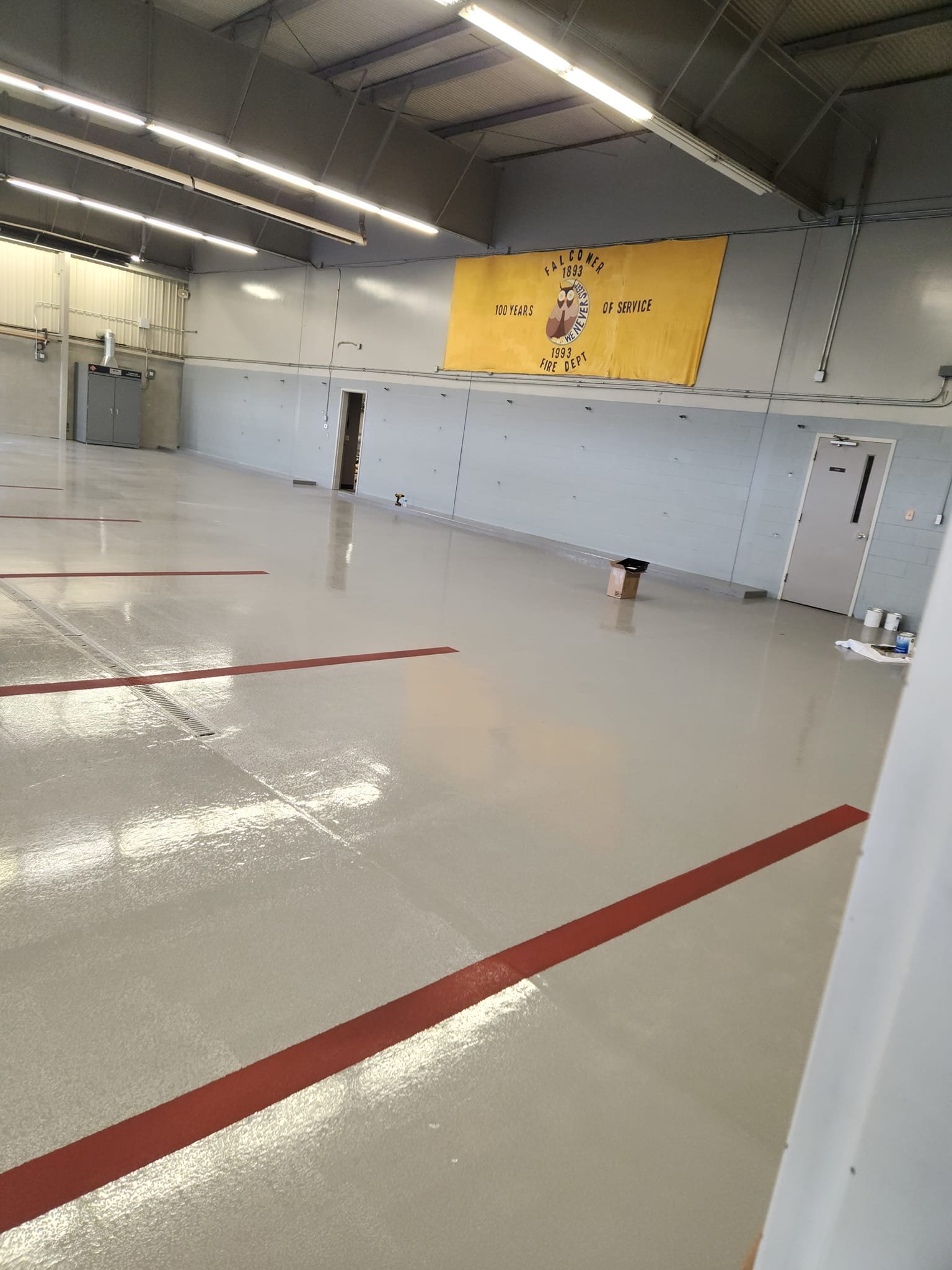Interior of an empty fire station garage with a banner on the wall celebrating 100 years of service for a fire department. The floor is polished and marked with red lines, and industrial lighting hangs from the ceiling.