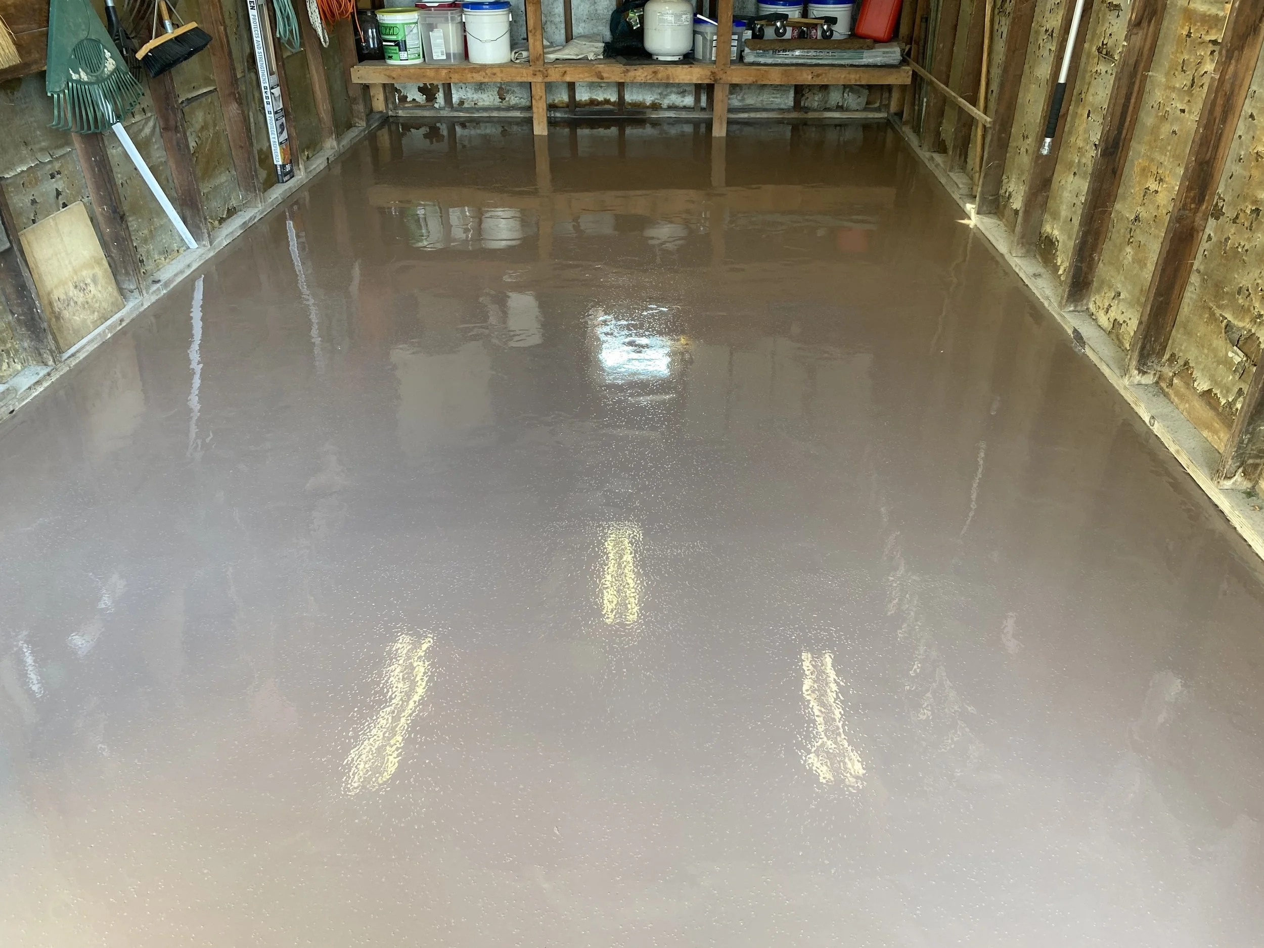 Freshly coated garage floor with a glossy finish, surrounded by wooden walls and various tools and supplies on shelves.