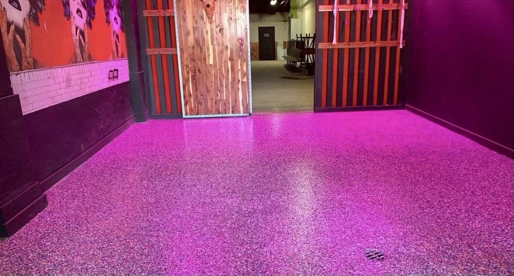 Room with a speckled pink floor, colorful mural on the left wall, and wooden doors.