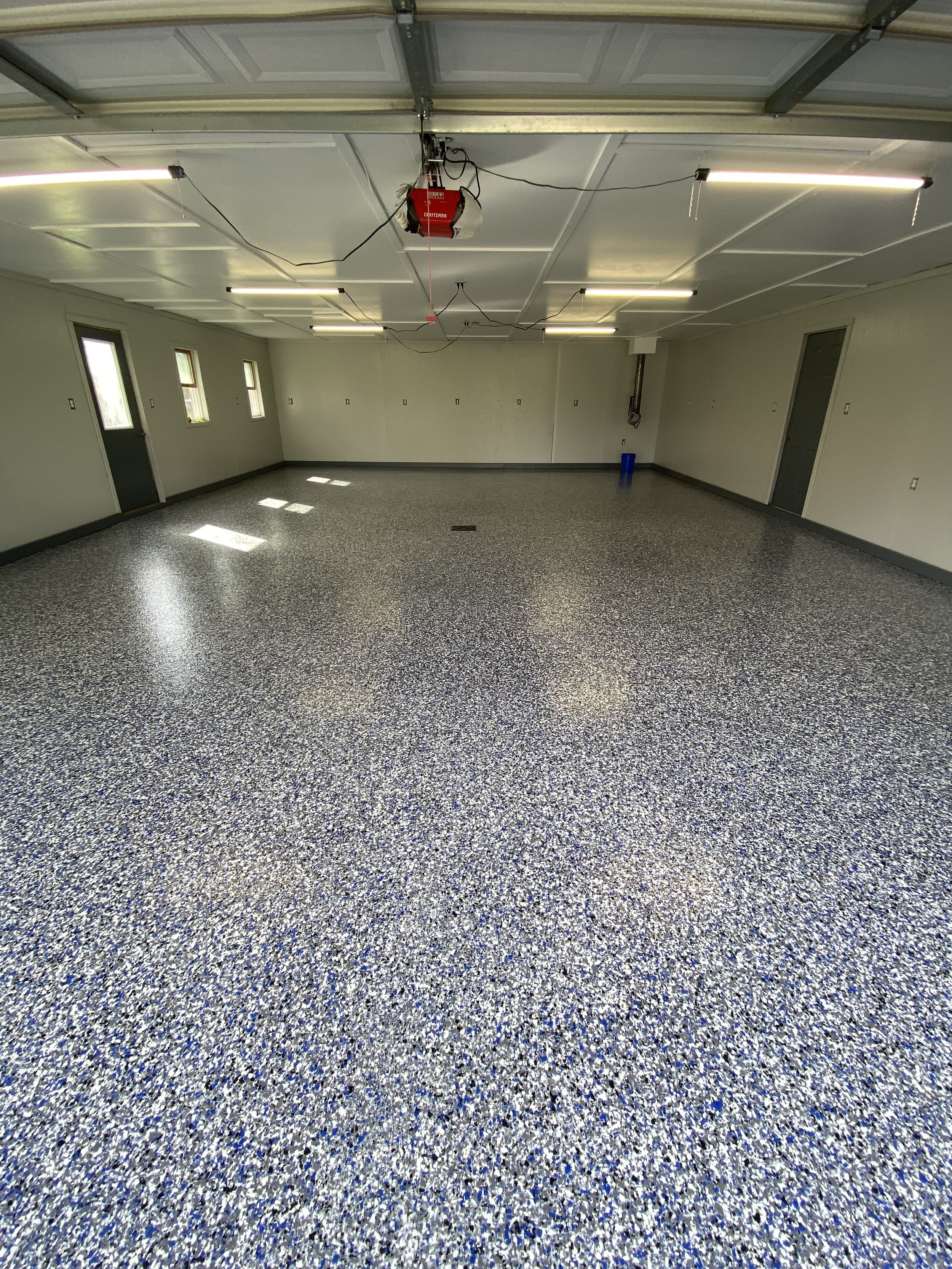 Empty garage with speckled epoxy flooring, white walls, ceiling lights, and a garage door opener.