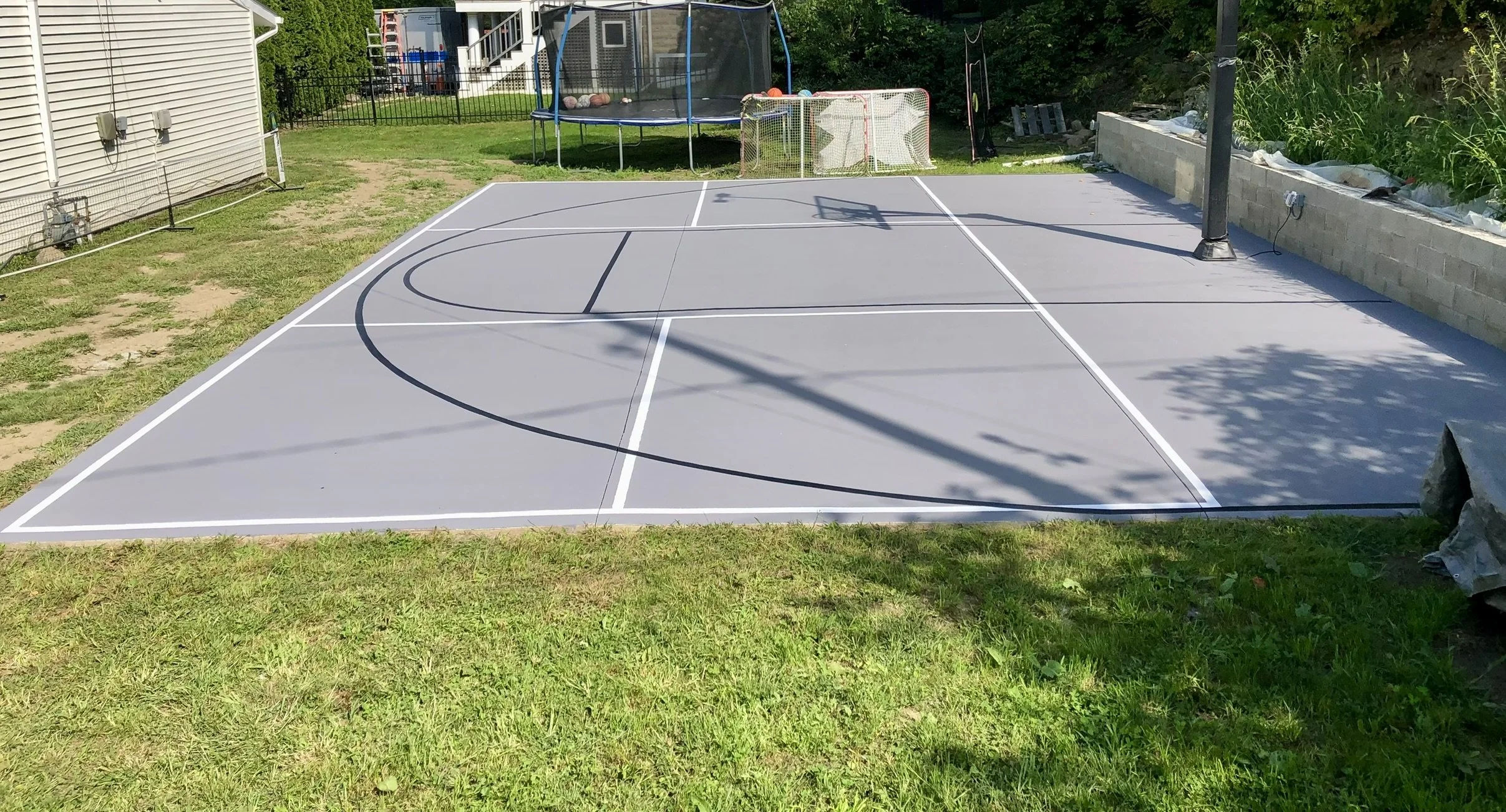 Outdoor basketball court in a backyard with a hoop, grass, netted trampoline, and hockey net in the background.