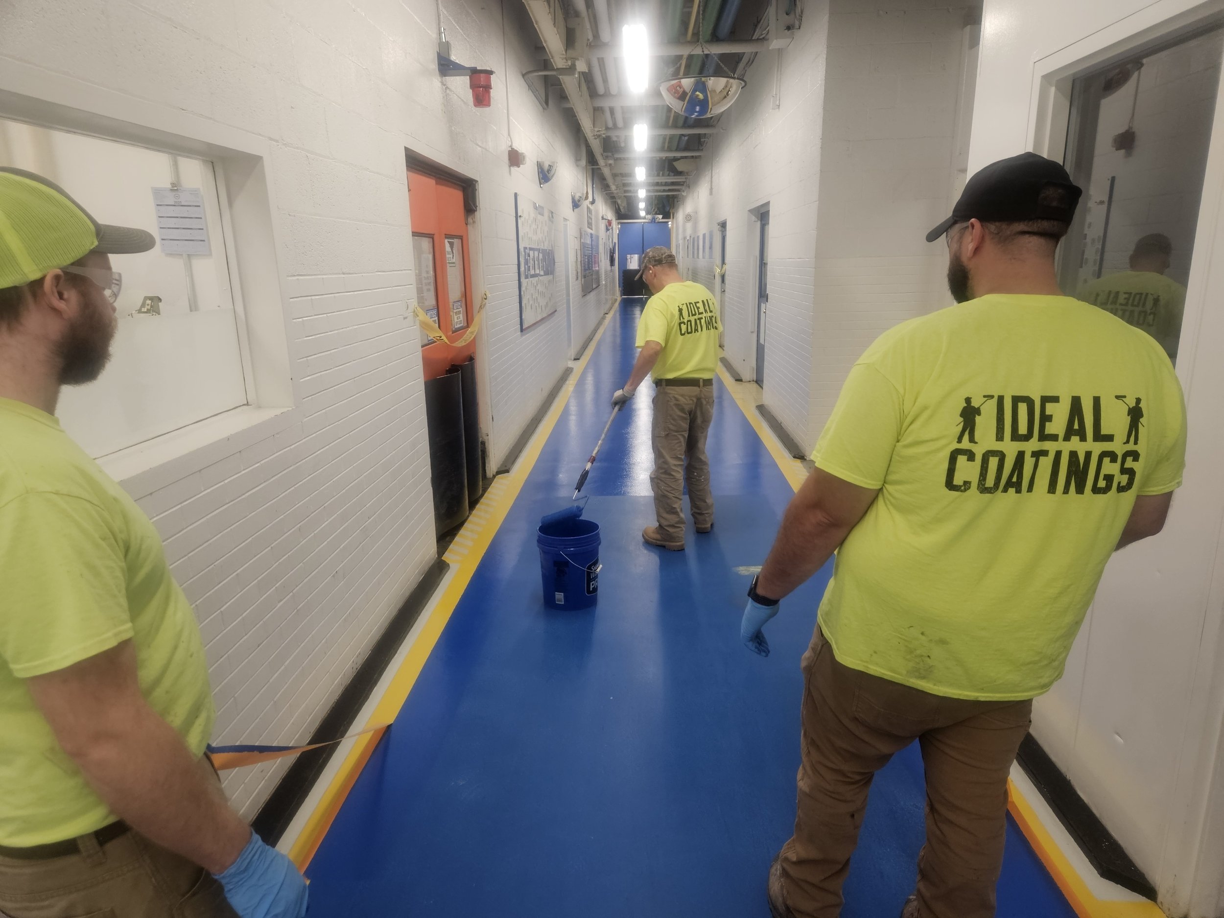 Workers in bright yellow shirts labeled "Ideal Coatings" apply blue coating to a hallway floor.