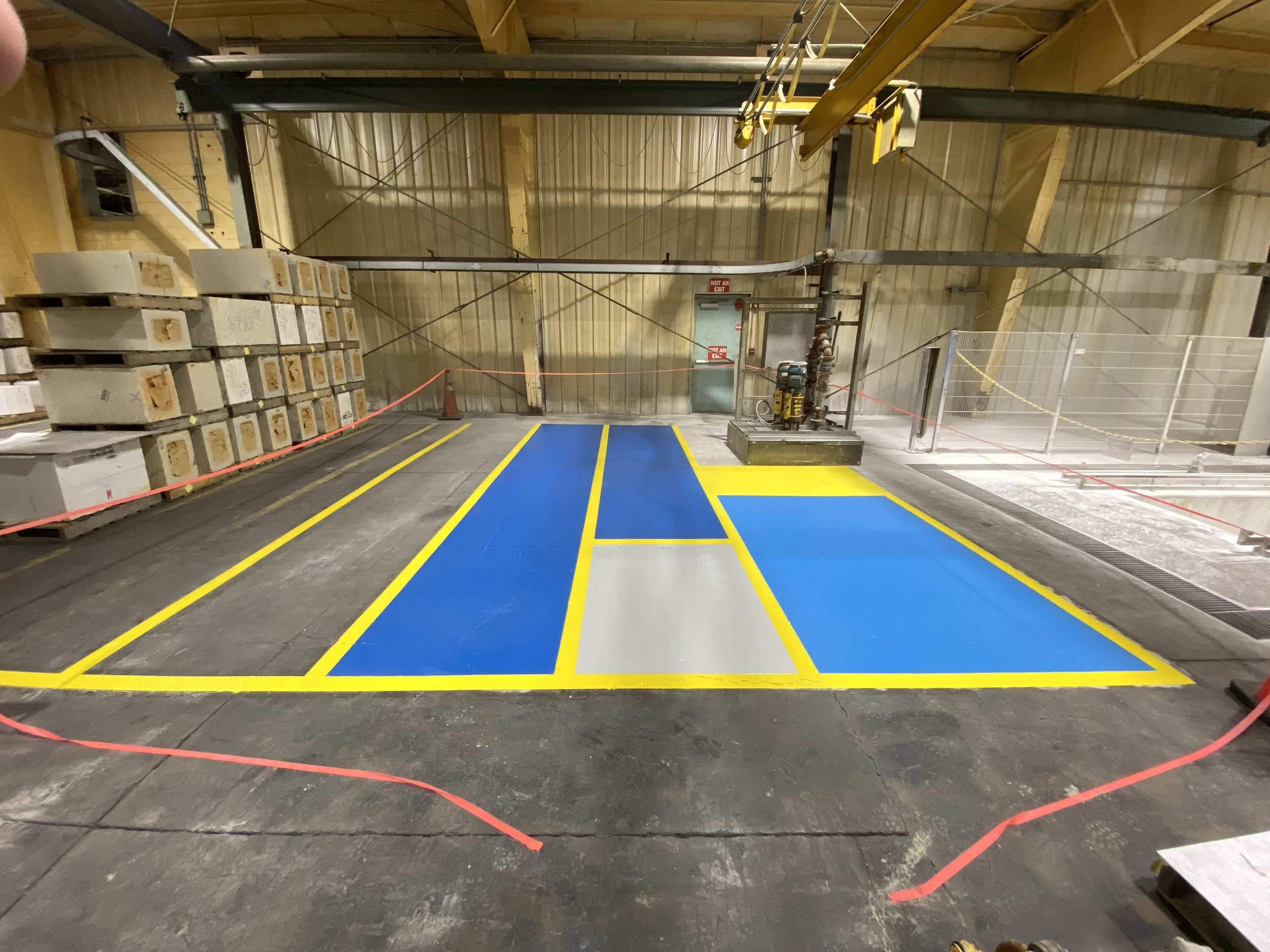 Industrial warehouse with painted safety zones on the floor, including blue and gray areas outlined in yellow, surrounded by stacks of boxes and machinery, with red caution tape.