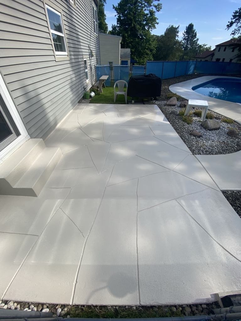 Backyard patio with light-colored paving stones, adjacent to a swimming pool, outdoor grill, and chairs. Fenced area with trees in the background.