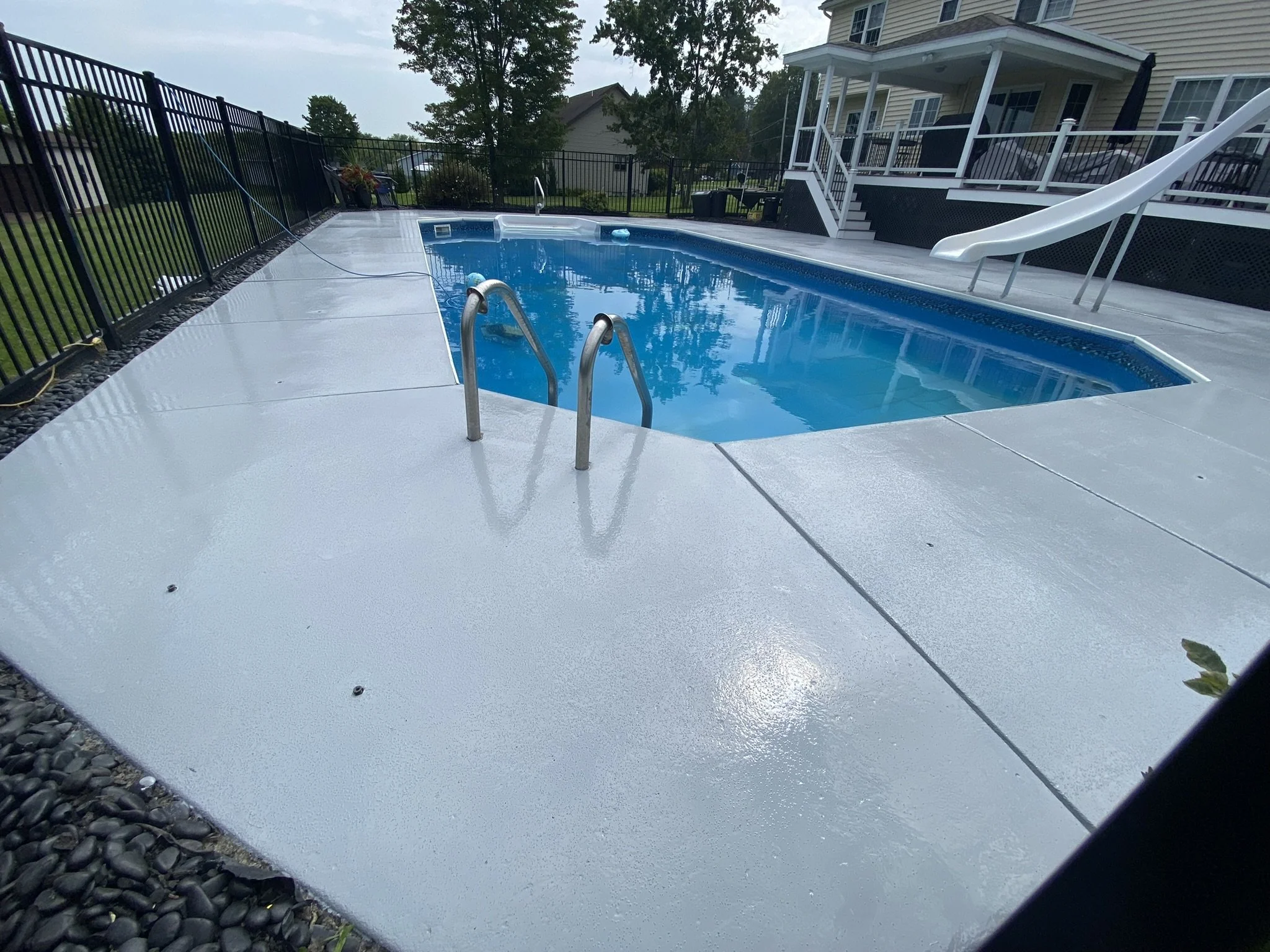 Backyard swimming pool with metal ladder and slide, surrounded by white concrete patio and fenced garden.