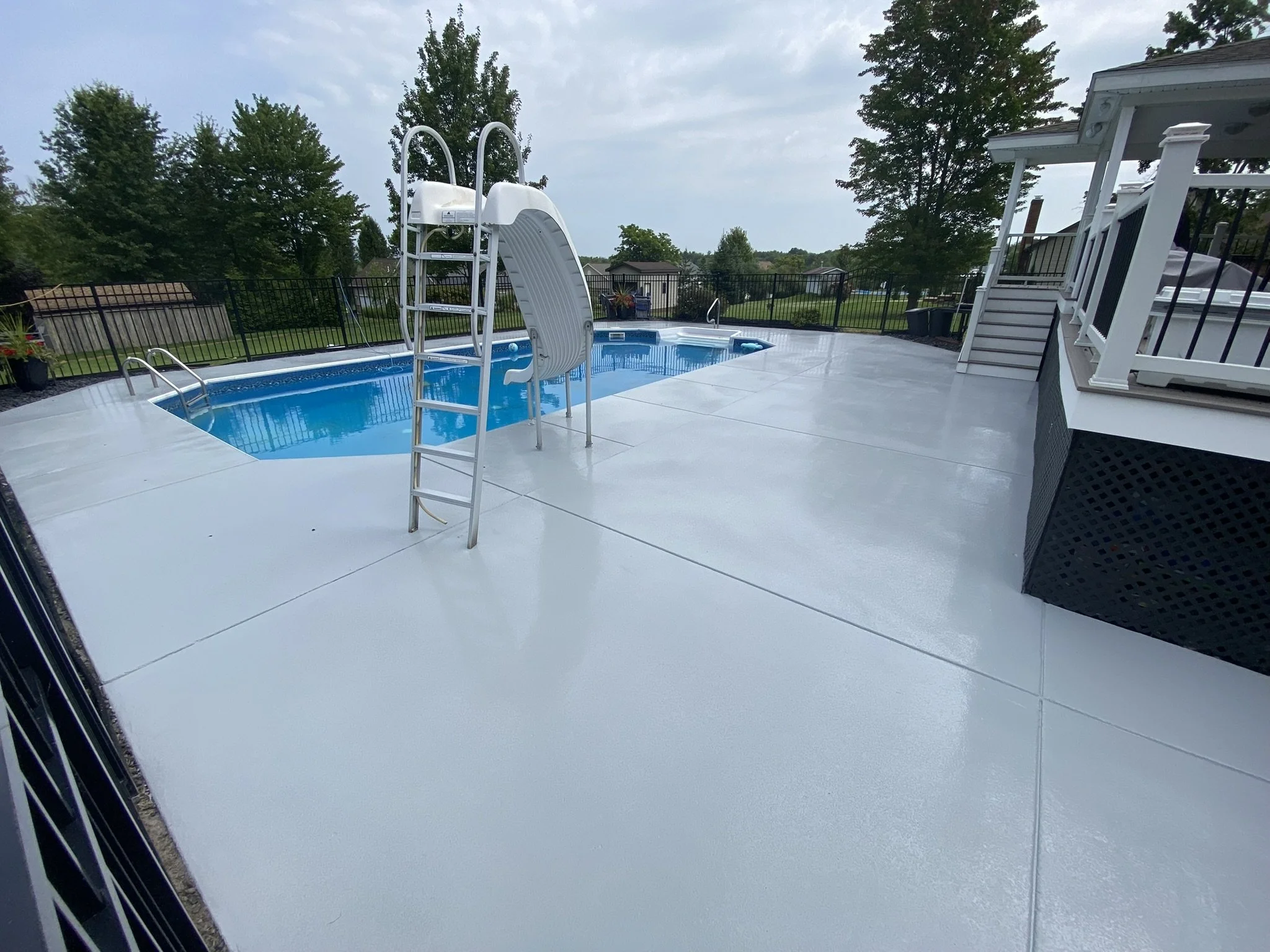 Backyard swimming pool with slide, surrounded by white concrete, trees in background.